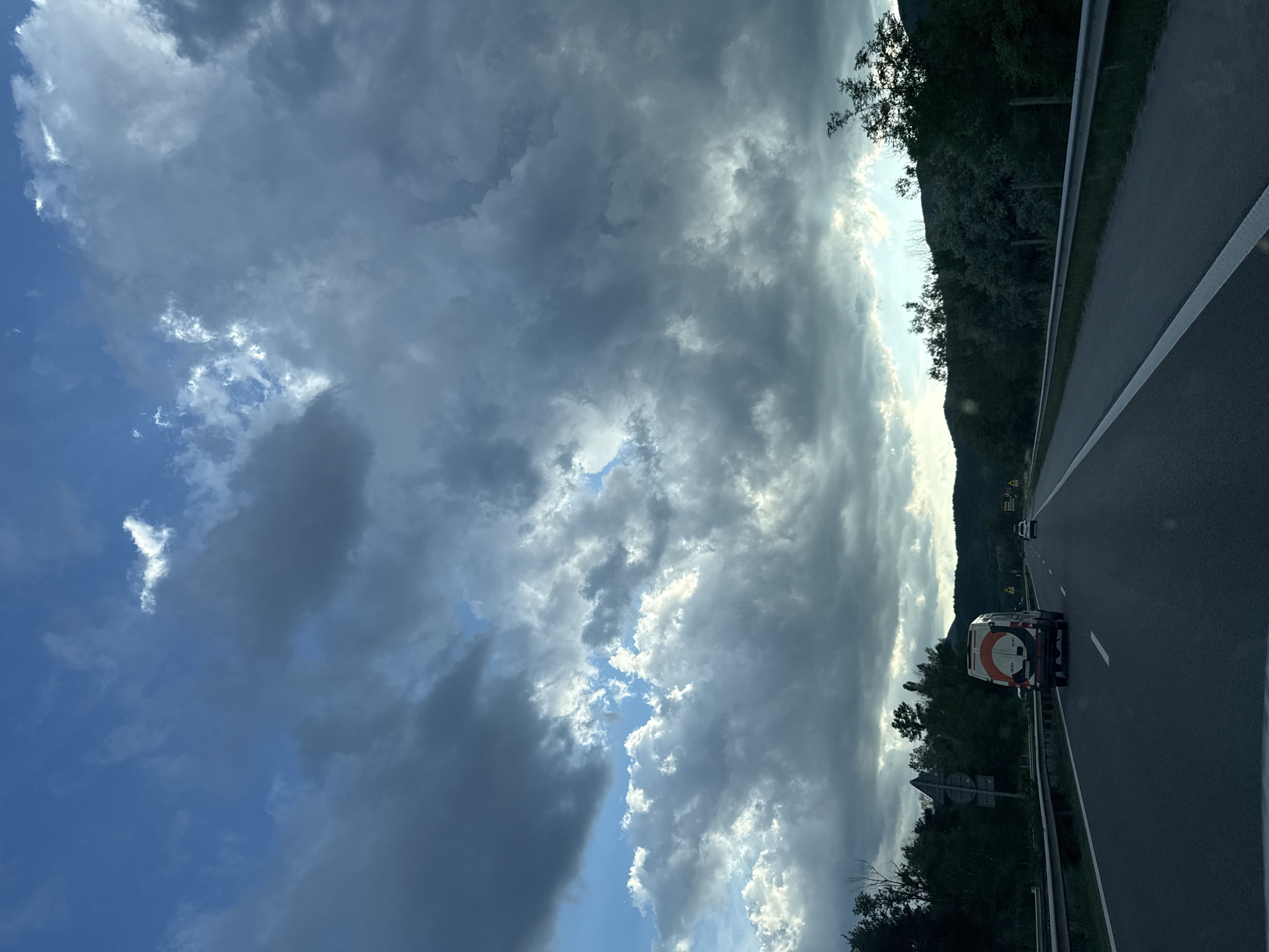 The image depicts a highway scene in the vicinity of Gorno Varshilo, Bulgaria. The sky is filled with dramatic, dark clouds interspersed with patches of bright blue sky and illuminated clouds, suggesting a dynamic weather condition. A multi-lane asphalt highway stretches into the distance, with white lane markings visible. In the foreground, a white van with a distinctive orange and black circular logo on its rear is driving in the right lane. Further ahead, a white passenger car is visible in the distance. The highway is bordered by a metal guardrail and lush green trees and vegetation lining the sides, with rolling hills in the background. The overall impression is one of travel through a scenic, somewhat overcast landscape.