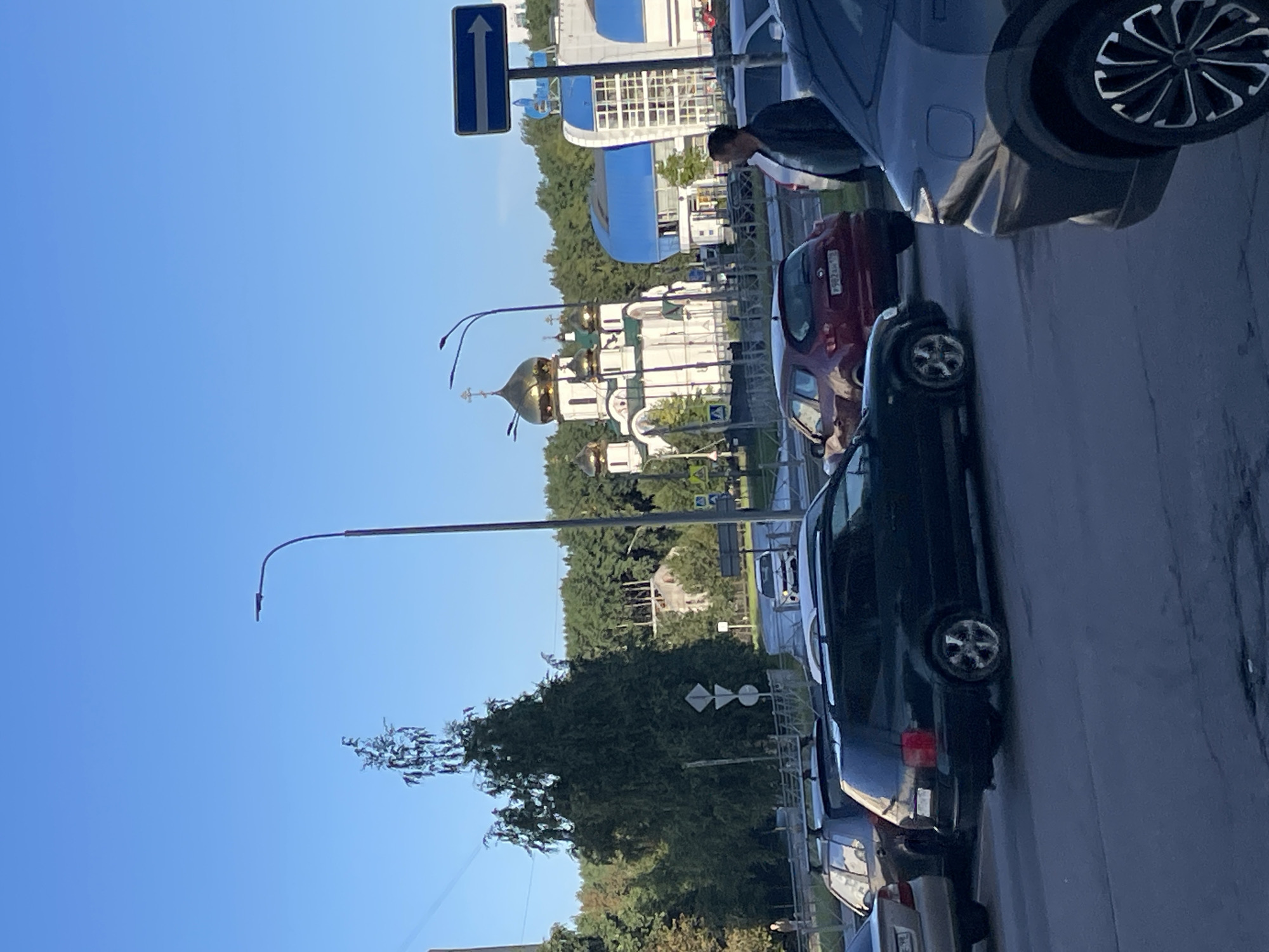 In a Saint Petersburg, Russia scene, a man stands near parked cars on a sunny day. In the background, a white Orthodox church with golden domes stands out, a modern building with a blue roof to its right. Trees line the horizon. Several cars are parked on the street, and others are driving.