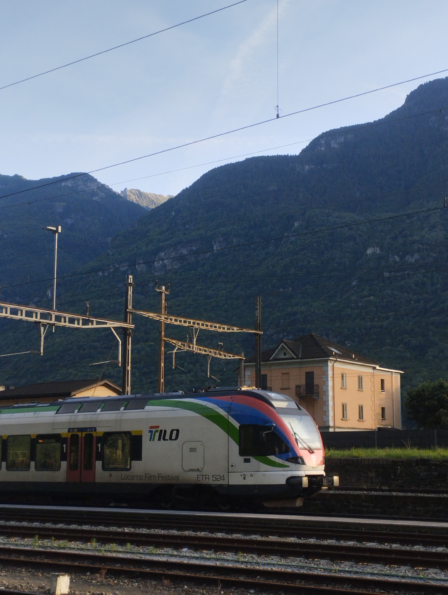 The image shows a train at a station with mountains in the background. The train is white with blue and green stripes and the logo "TILO" is visible on its side. The words "Locarno Film Festival" and "ETR 524" are also visible on the train. There is a light yellow building with a dark roof behind the train, and railway tracks in the foreground. The sky is blue and clear, and the mountains are covered in green trees. The scene appears to be a daytime shot, possibly in the morning or late afternoon, given the lighting. There are no people visible in the image. The overall impression is one of a scenic train journey in a mountainous region.