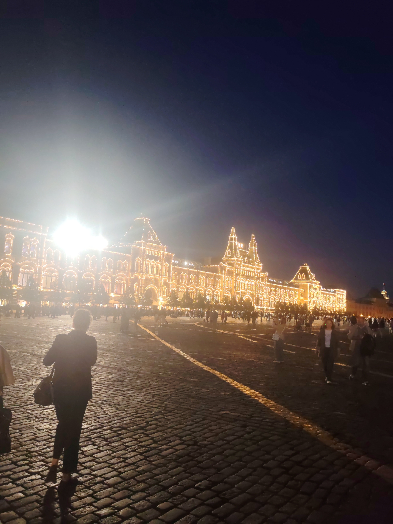 In an outdoor, cobblestone square, a large, ornate building is illuminated by numerous strings of warm, yellow lights that outline its architectural features. The building has a grand facade with arched windows and multiple towers, and it appears to be a historic structure. A bright light source, possibly a spotlight or the moon obscured by haze, is visible in the upper left portion of the frame, creating a strong glare and obscuring some details.
Several people are present in the square, walking and standing. In the foreground, a person is captured from behind, wearing a dark jacket and dark pants, carrying a shoulder bag. Their silhouette is distinct against the paved ground. Other individuals and groups are scattered throughout the midground and background, their forms less defined due to the lighting and distance. The sky is a deep, dark blue, suggesting it is nighttime or twilight. The overall atmosphere is festive, likely due to the elaborate lighting display on the building.