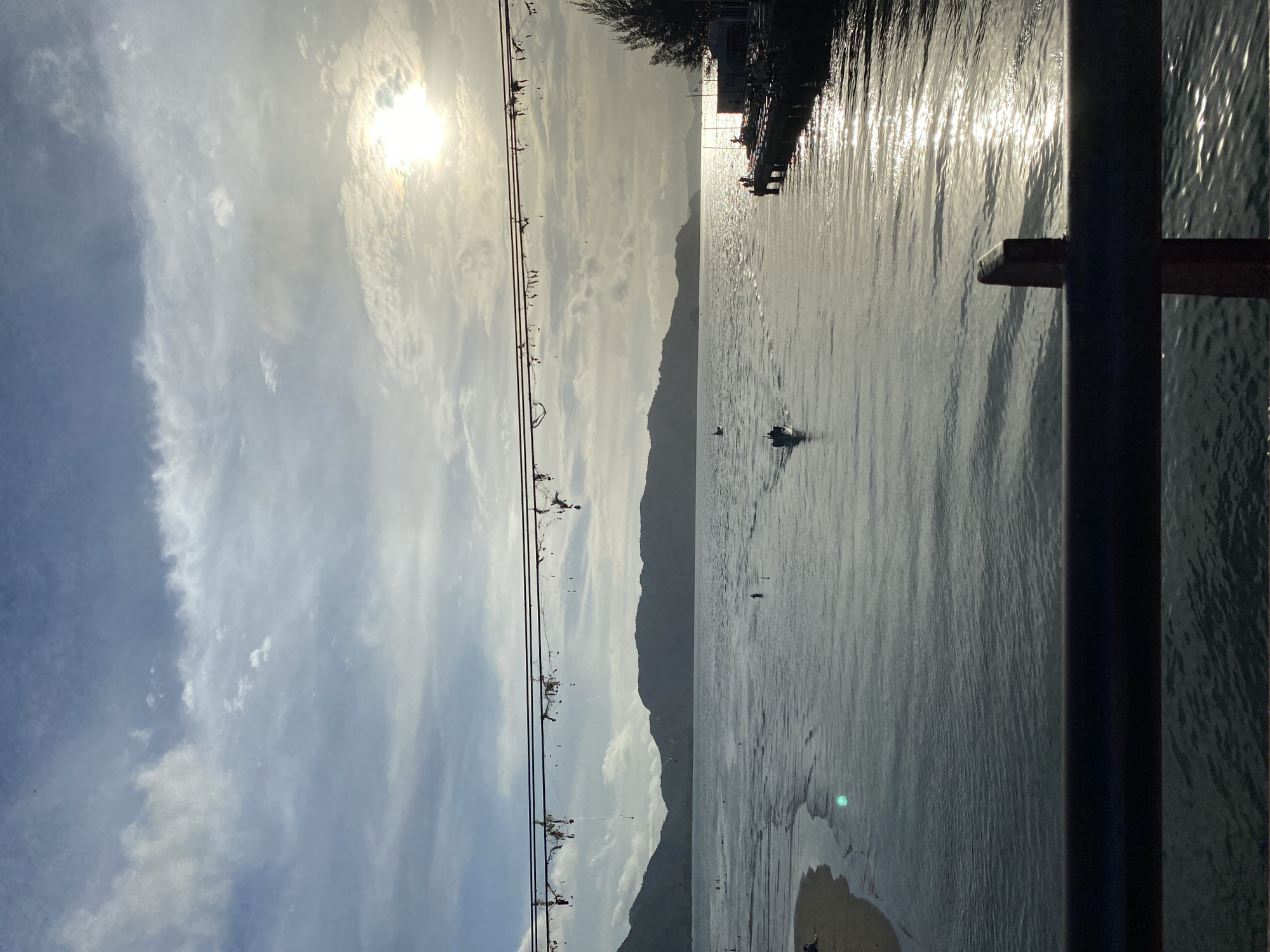 This image captures a serene coastal scene in Banda Aceh, Indonesia, likely during late afternoon or early morning given the low angle of the sun. The foreground features a dark railing, suggesting the viewpoint is from a pier, bridge, or elevated walkway. Beyond the railing, a body of calm water stretches towards a mountainous horizon. The water is reflecting the bright, hazy sunlight, creating shimmering patterns on its surface.
In the middle ground, the calm sea is dotted with a few small boats and several figures. One boat is moving, leaving a subtle wake, while another appears stationary. Several individuals are visible in the shallow water near the shore on the left, and one person is standing in the water further out. On the right side of the frame, a wooden pier extends into the water, with a couple of figures standing on it. A building, possibly a restaurant or shop, is situated at the end of the pier.
The sky above is a mix of blue and white clouds, with the sun partially obscured by a layer of thin clouds, creating a bright, diffused light. The distant hills are silhouetted against the sky. Overhead, a series of wires, likely power or communication lines, are strung across the frame, with some decorative elements or perhaps remnants of decorations hanging from them. The overall impression is one of tranquility and natural beauty.