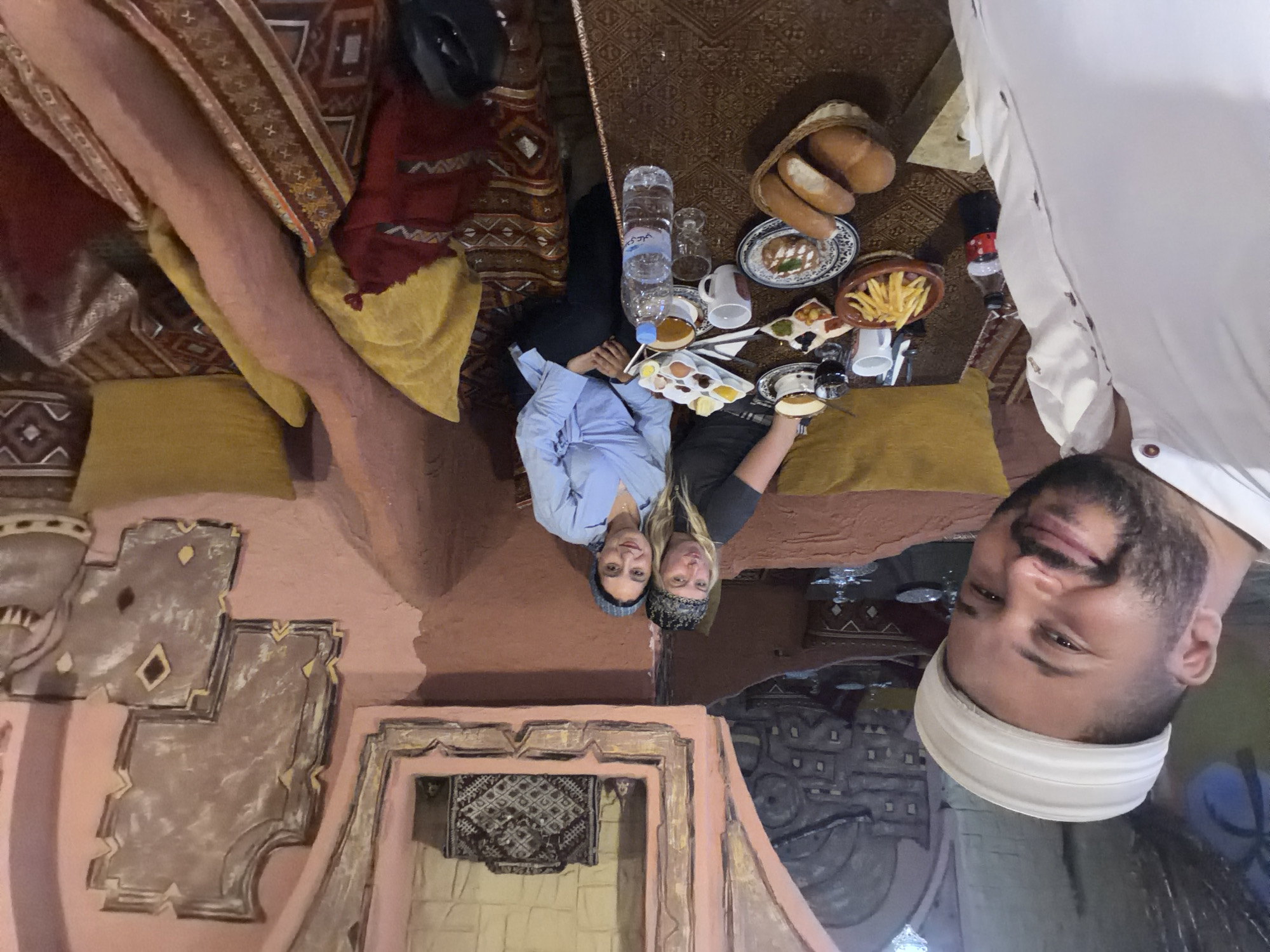 The image captures a group of three people enjoying a meal in what appears to be a traditional Moroccan setting, likely a restaurant or riad in Kenitra, Morocco. The photo is taken from a low angle, possibly a selfie.
In the foreground, a man with a beard and a white headwrap or turban is visible, smiling slightly, facing the camera. Behind him, two women are seated on patterned cushions on the floor, facing the viewer and also smiling. One woman has dark hair and wears a blue long-sleeved shirt with a dark head covering. The other woman has blonde hair and a patterned head covering over a dark top.
Between the women is a low, intricately patterned table laden with various food items. These include a basket of bread, multiple small plates with what appear to be breakfast or brunch dishes such as eggs, spreads, and pastries. A plate of french fries, cups of coffee or tea, and a large bottle of water with Arabic text ("سيدي علي" - Sidi Ali, a Moroccan brand) are also visible. The floor is covered with ornate, earthy-toned rugs. The walls feature decorative architectural reliefs, painted in pinkish-brown and beige tones, adding to the traditional ambiance. The overall mood is relaxed and joyful. The interior lighting suggests an indoor setting, possibly during the morning or early afternoon given the breakfast-like spread.