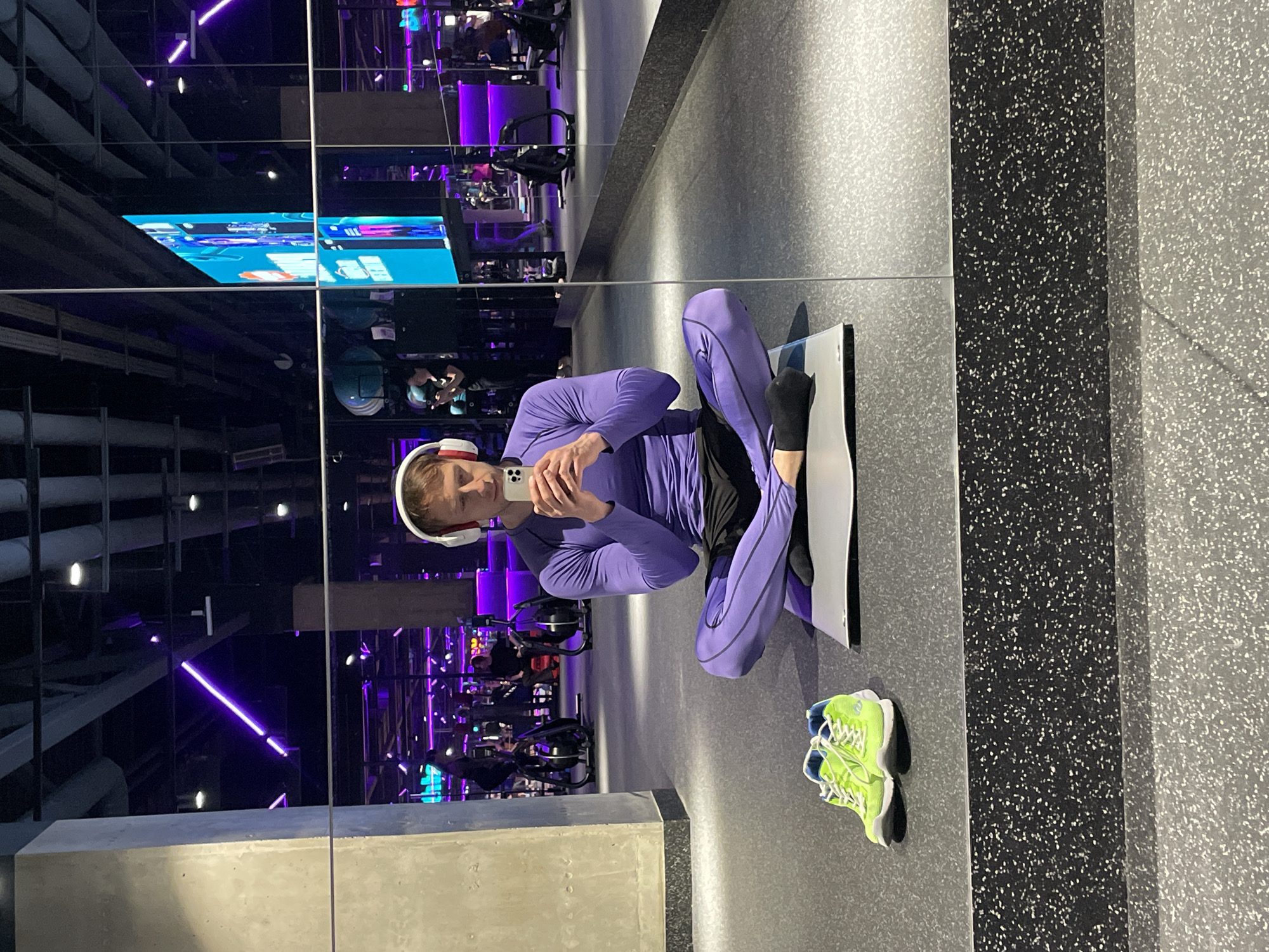 A mirrored view shows a person in purple athletic wear and headphones, holding a phone and seemingly taking a selfie while kneeling on a white yoga mat. Neon green shoes are near the mat. The backdrop includes exercise equipment and purple neon lighting. The location appears to be a gym. The city of Murino, Russia is not discernible.