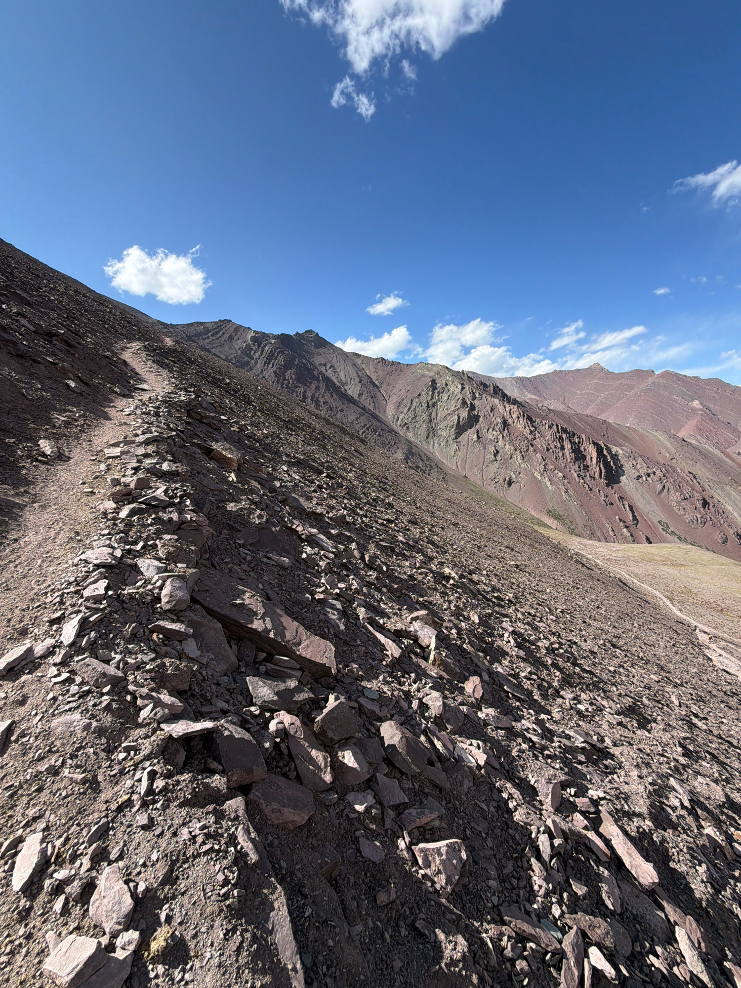 A steep, rocky mountain slope in a remote, high-altitude location. The path, made of dirt and loose stones, winds its way up the mountainside. The mountains in the background are rugged and bare, with reddish-brown and grey rock formations. The sky is a vibrant blue with scattered white clouds. The overall impression is one of a wild and desolate landscape.