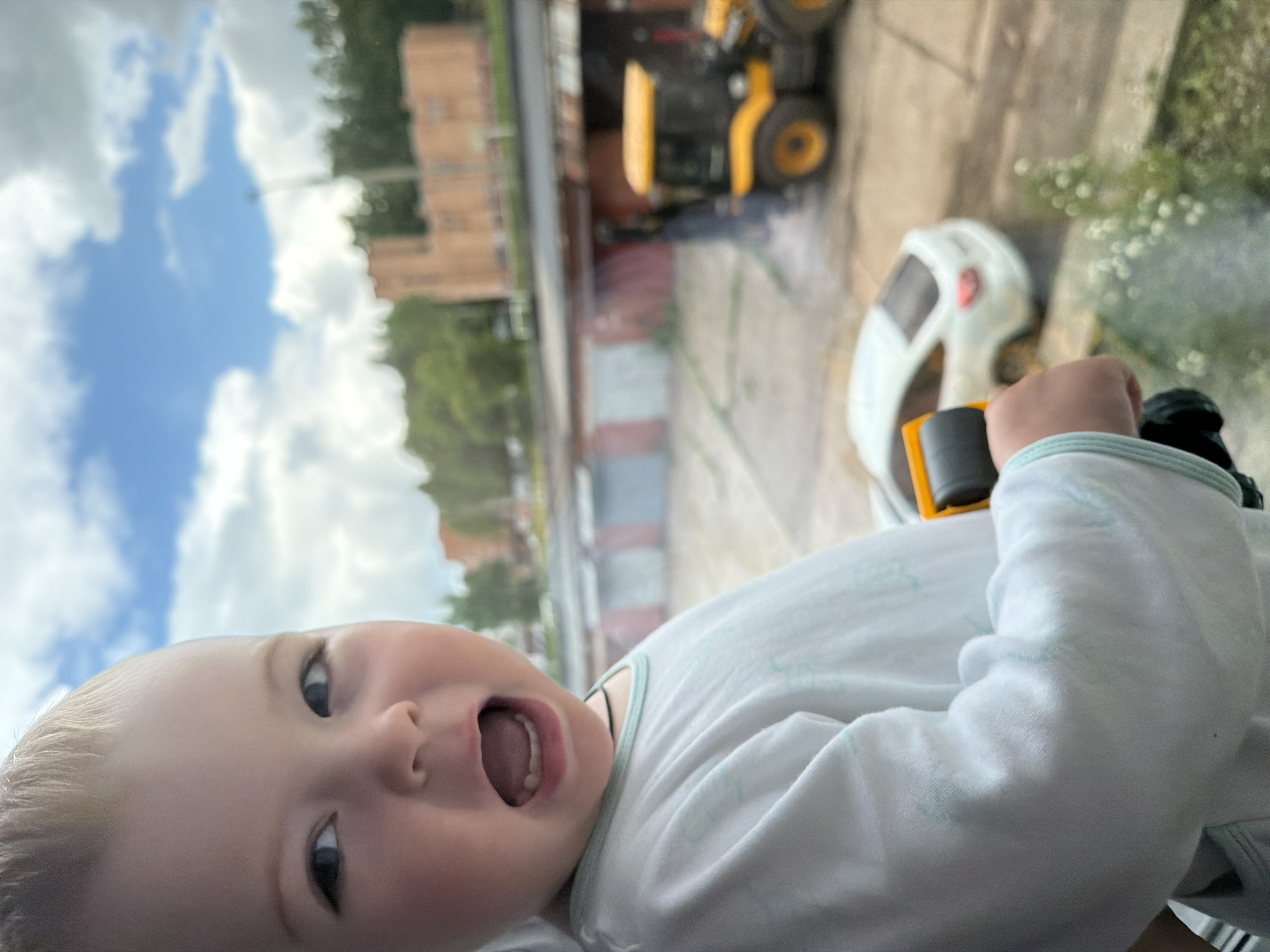 This sideways-oriented image captures a joyful young child with blonde hair and blue eyes, looking directly at the camera with an open, smiling mouth. The child wears a light-colored top with a light blue trim and holds a small white toy vehicle with yellow accents.
The background, seen through what appears to be a window, is out of focus but provides context. A bright blue sky with fluffy white clouds dominates the upper left. Below this, blurred green trees and red-brick buildings suggest an outdoor, semi-urban setting in Votkinsk, Russia. On the right side of the image, two blurred vehicles are visible: a larger yellow construction vehicle, possibly a backhoe or excavator, and a smaller white vehicle resembling a toy car, parked on paved ground with some greenery.
The activity shows the child observing the outside world, possibly playing with a toy that mirrors the vehicles visible through the window. The bright lighting indicates daytime, under sunny conditions with scattered clouds. The child's wide eyes and open mouth convey a sense of happiness, excitement, or surprise. There is no visible text in the image.