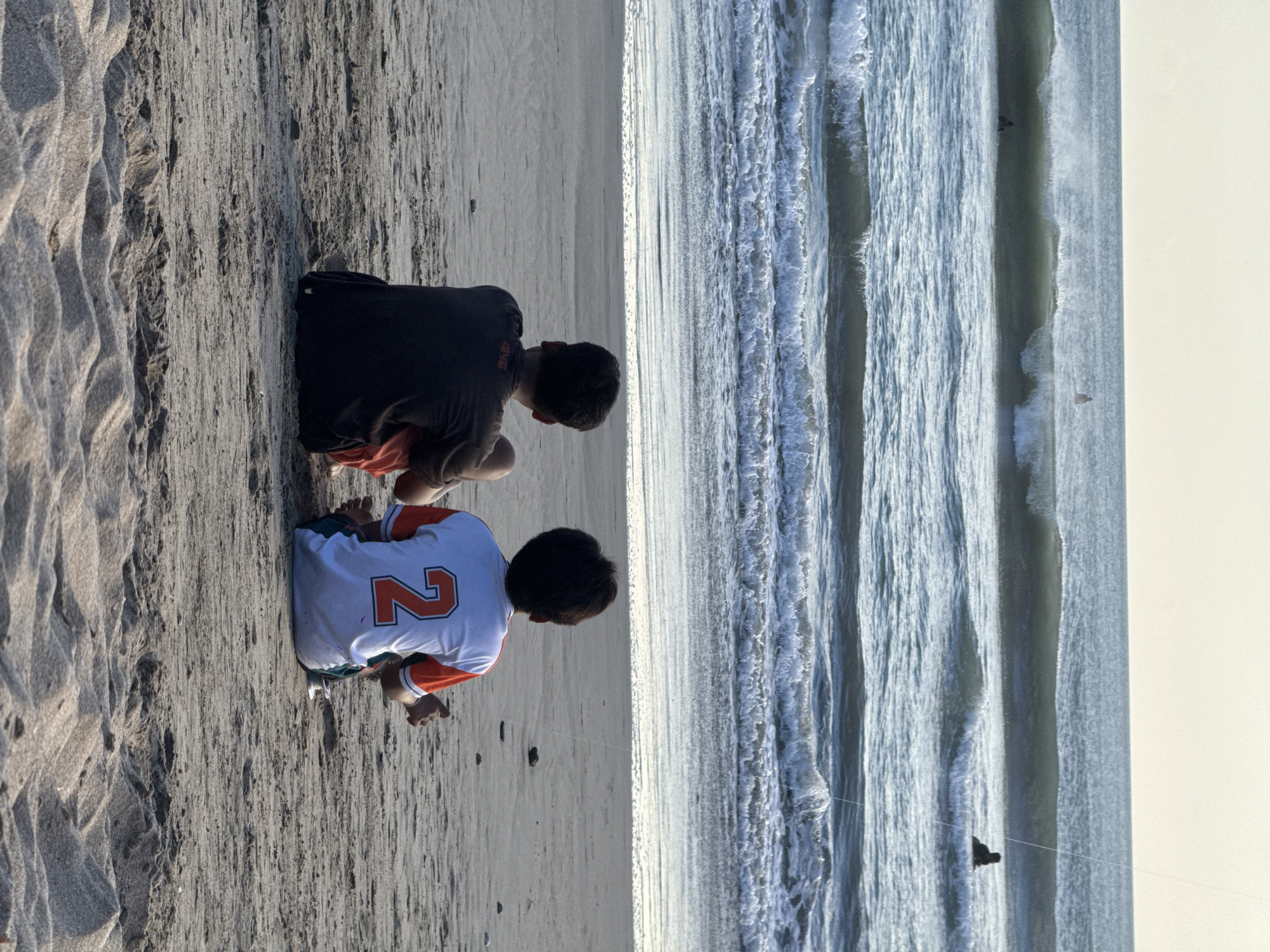 Two young males are sitting on a sandy beach in Kuta, Indonesia, facing the ocean. The male on the left wears a dark t-shirt and orange shorts. The male on the right wears a white t-shirt with an orange collar and sleeves, featuring a large orange number "2" on the back. His shorts appear to be green. Both are seated with their legs crossed or tucked beneath them, looking out at the sea.
The ocean in front of them has several sets of waves rolling in, with white foam cresting on the closer waves. In the distance, the sea is calmer, and a few individuals are visible in the water, appearing to be swimming or surfing. The sky above is a pale, light color, suggesting either early morning or late afternoon light. The sand on the beach is light gray and textured with small ripples and a few scattered pebbles. The foreground shows more prominent sand ripples, indicating wind movement or water action.