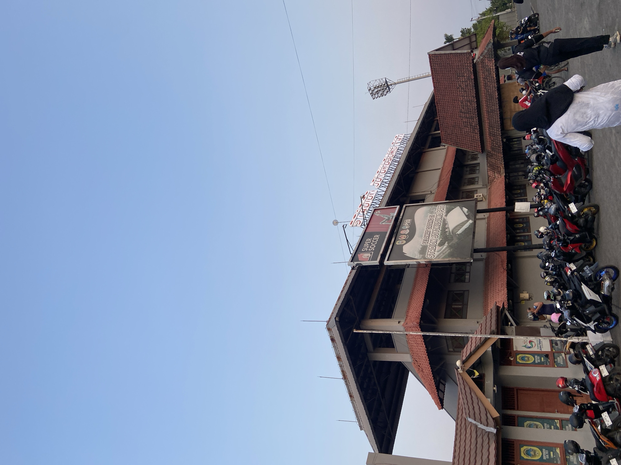 The image depicts the exterior of a stadium in Margorejo, Indonesia, under a clear blue sky. The stadium features a multi-tiered structure with red tiled roofs and gray walls. Prominently displayed on the upper level is signage that reads "STADION JATUKUSUMO PATI" and "SUPER SOCCER". A tall floodlight tower is visible to the right of the stadium. In front of the stadium, a large number of motorcycles are parked, suggesting it is a public venue. Several individuals are present, some walking and others near the parked motorcycles. The overall scene is one of an outdoor sports facility in an urban or semi-urban setting.