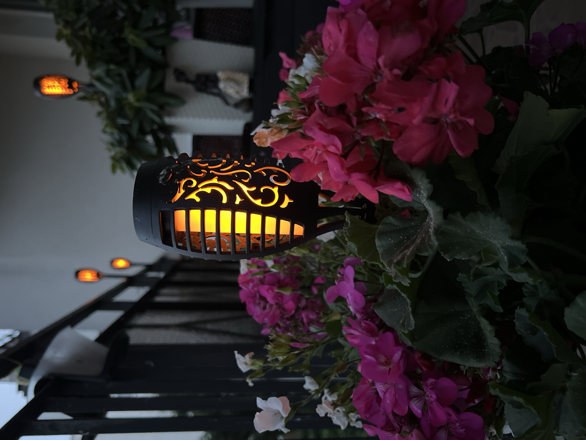 The image depicts a close-up view of a solar-powered torch light with a flickering flame effect, situated amidst a cluster of bright pink and white flowers in a planter. The torch light has intricate, ornamental cutouts on its casing, allowing the warm, orange light to glow through. In the background, a dark metal railing is visible, with other similar torch lights spaced along it, illuminating a balcony or patio area. Beyond the railing, a light-colored wall and some greenery are present. The overall setting appears to be an outdoor space, likely a balcony or garden, in the city of Kleosin, Poland, during twilight hours. No people are visible in the frame.