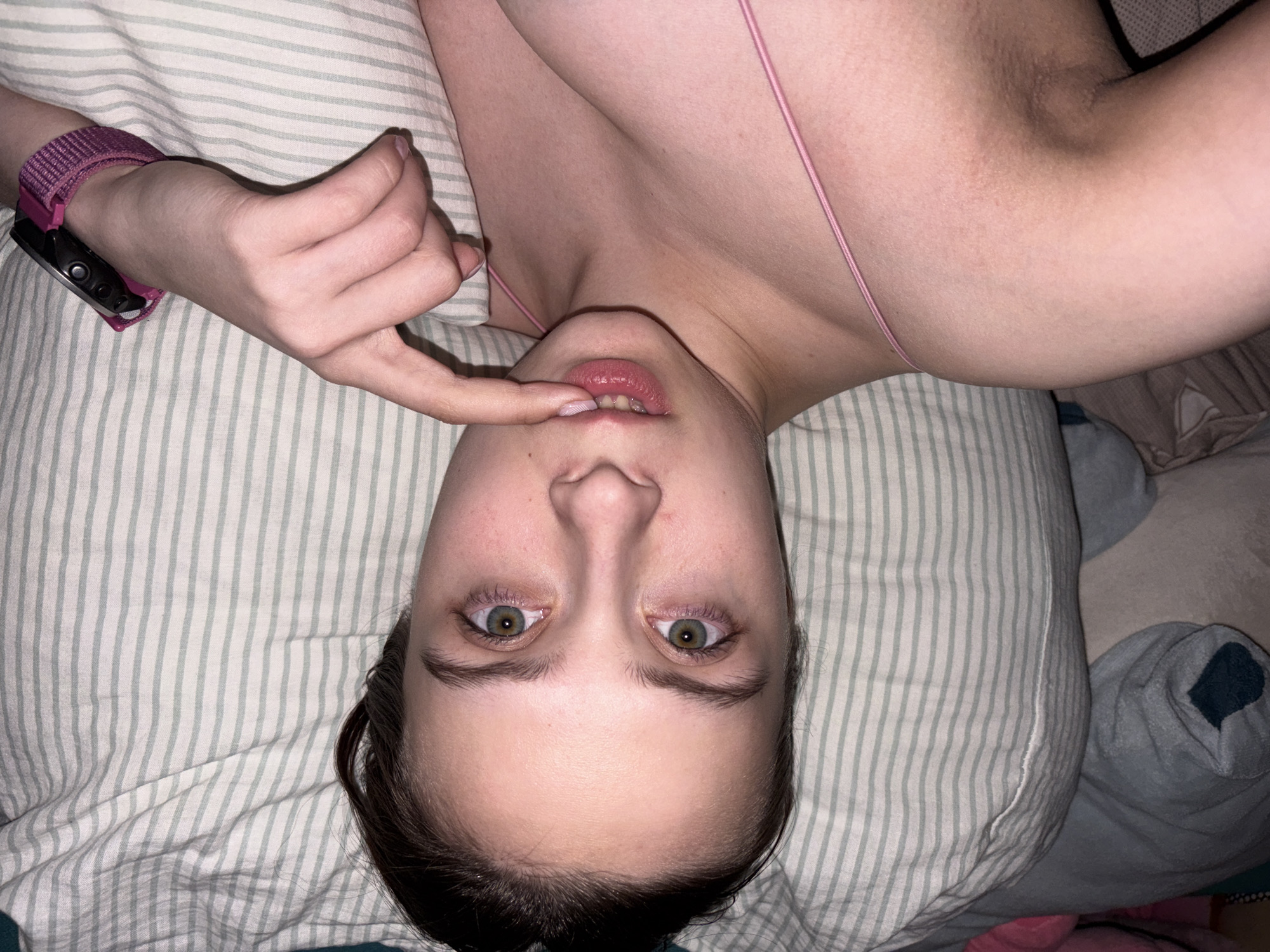 The image captures a close-up, overhead view of a young person lying down, with their head positioned upside down in the frame. The subject has fair skin, dark hair, and striking green-hazel eyes that are wide open and looking directly at the viewer. Their lips are slightly parted, revealing some lower teeth, and their right index finger is gently touching their lower lip.
The person is wearing a thin, pink strap, likely part of a bra or camisole, visible across their chest and shoulder area. On their left wrist, a watch with a vibrant pink fabric strap and a black watch face is visible. The background is dominated by a light-colored pillow with thin, light green vertical stripes, suggesting a bed or lounge setting. To the right, a soft, light blue textured object, possibly another pillow or blanket, is partially visible. The lighting is soft and appears to be indoors, possibly in low light conditions such as evening or night. No text is visible within the image. The provided location context for this image is Прикамский, Russia. The expression is one of direct engagement or thoughtful contemplation.