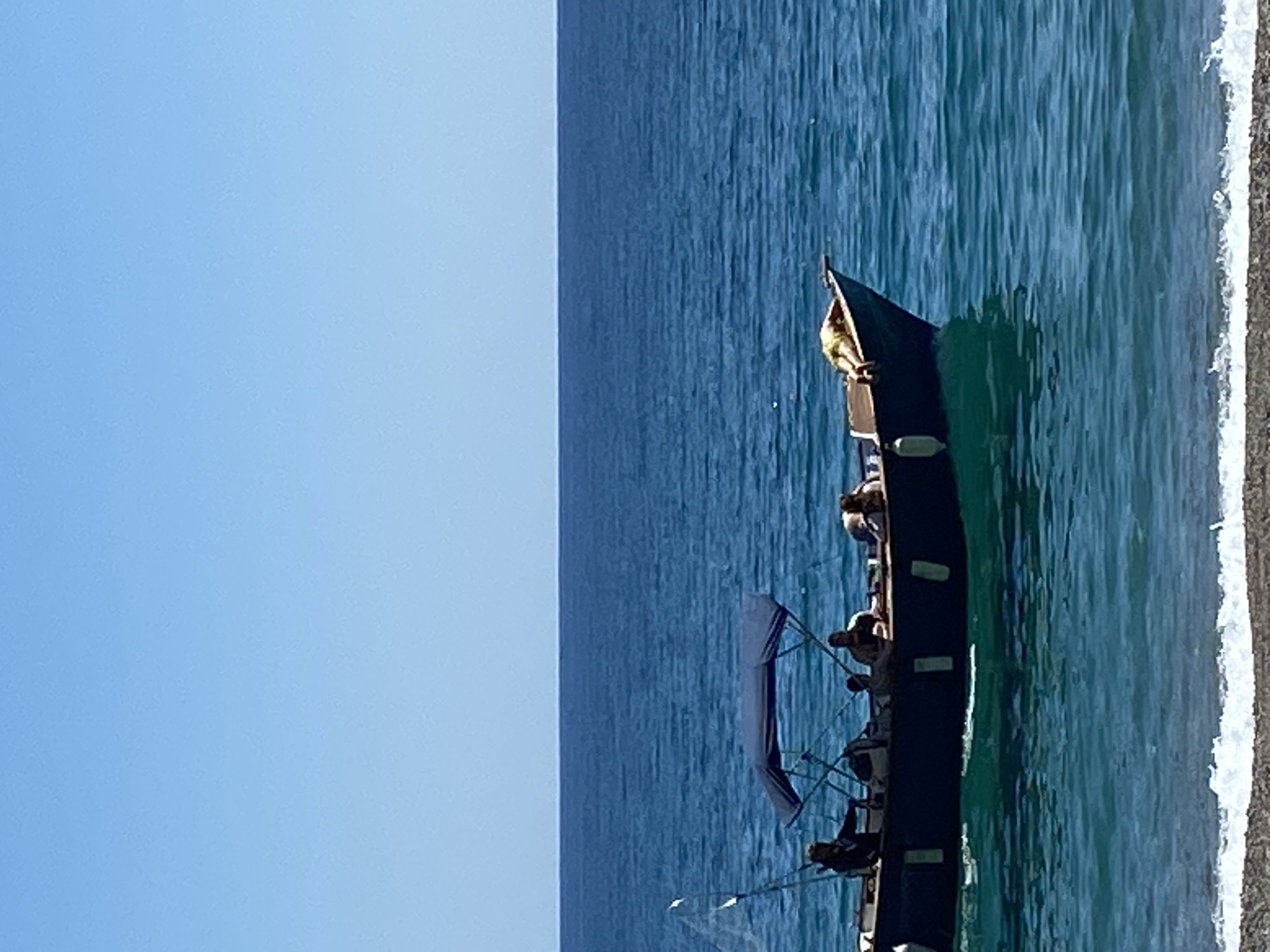 The image depicts a long, narrow boat, possibly a tour boat or large gondola, positioned close to the shore of a body of water. Several individuals are visible on the boat. The boat is surrounded by water that exhibits a light teal coloration. The shore consists of a light gray sandy or pebble-covered surface. The background is split between a clear, light blue sky and the dark blue of the sea stretching to the horizon. The image is possibly located in Prijevor, Montenegro, however this is unconfirmed from the image alone.