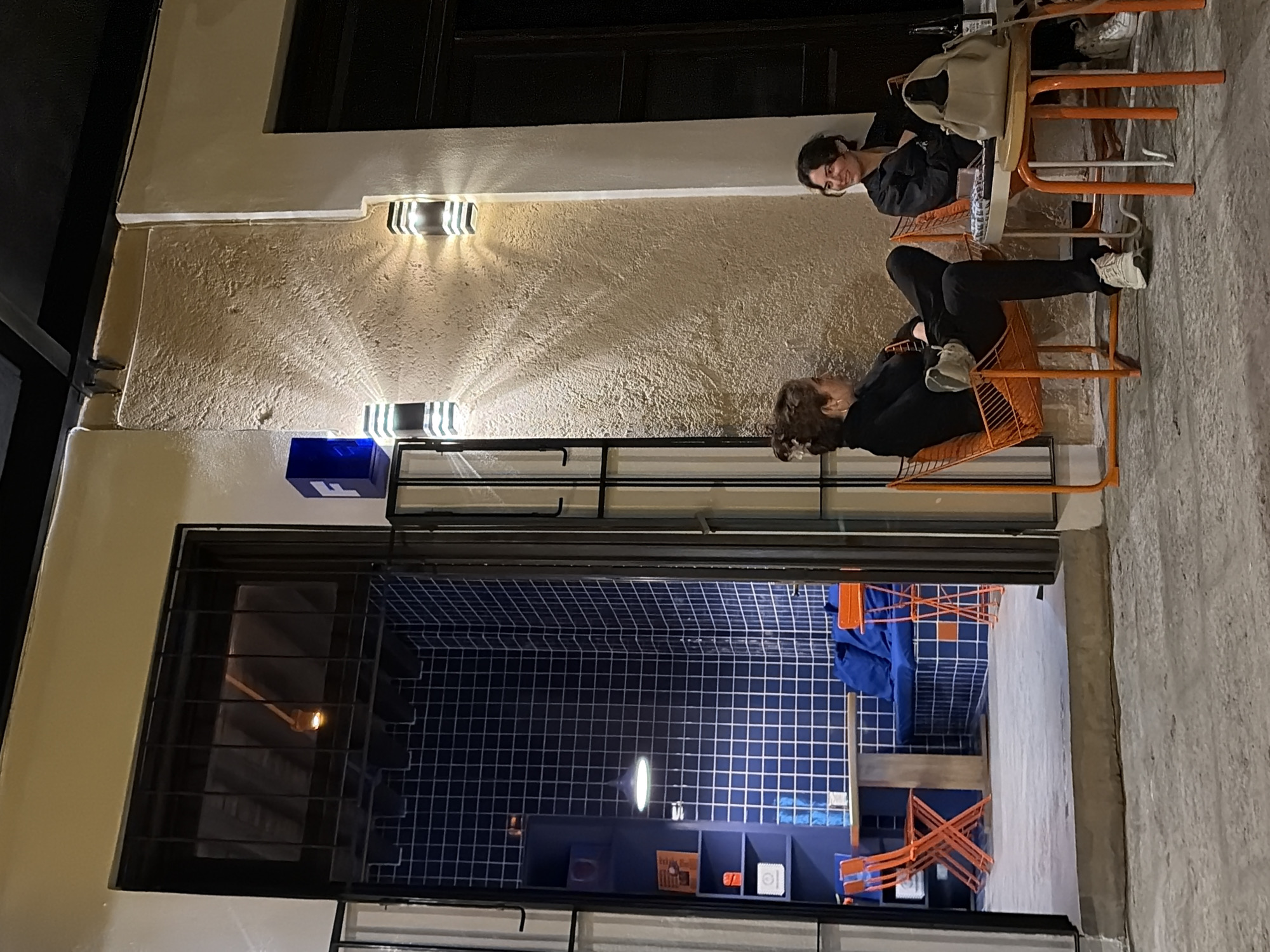 Two women sit on orange chairs outside a building in Puebla, Mexico. The building has an open window with a blue tiled interior. Exterior lights illuminate a stucco wall. A blue box is above the window. One woman faces the viewer while the other leans back.
