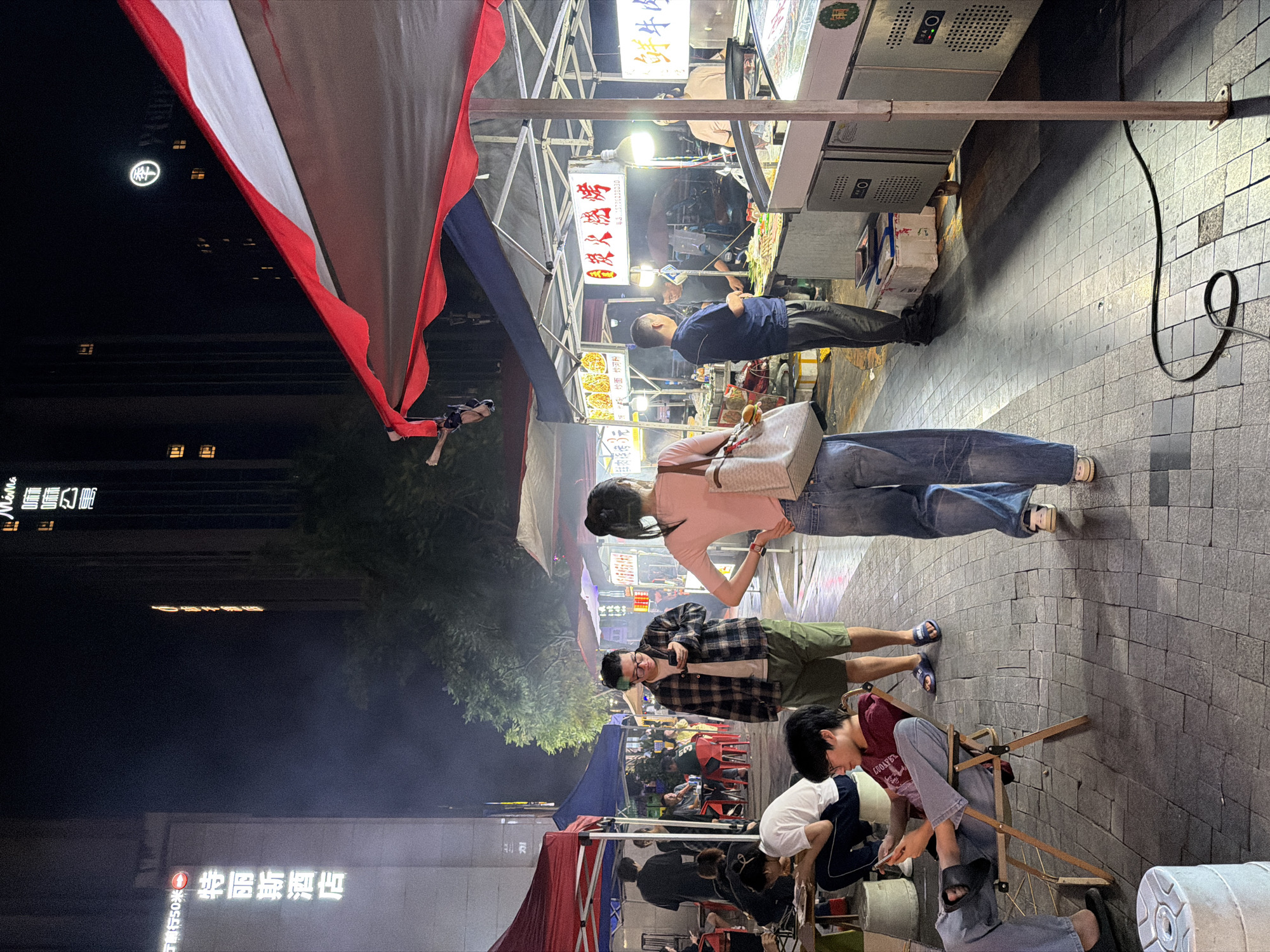 At night, a bustling street market is depicted in the Chongqing High-tech Zone, China. In the foreground, a woman in a pink t-shirt and blue jeans walks away from the camera, carrying a large tote bag. To her left, a man in a plaid shirt and shorts is looking at his phone. Further left, two individuals are seated at a table under a red tent, with one person eating.
The market features several food stalls with brightly lit signs and awnings. One stall displays a variety of skewers and grilled food, with vendors preparing and serving customers. Another stall appears to be selling seafood. Smoke or steam rises from some of the cooking areas, adding to the atmosphere of the busy night market. Tall buildings, some illuminated, are visible in the background, indicating an urban setting. The ground is paved with stone tiles.