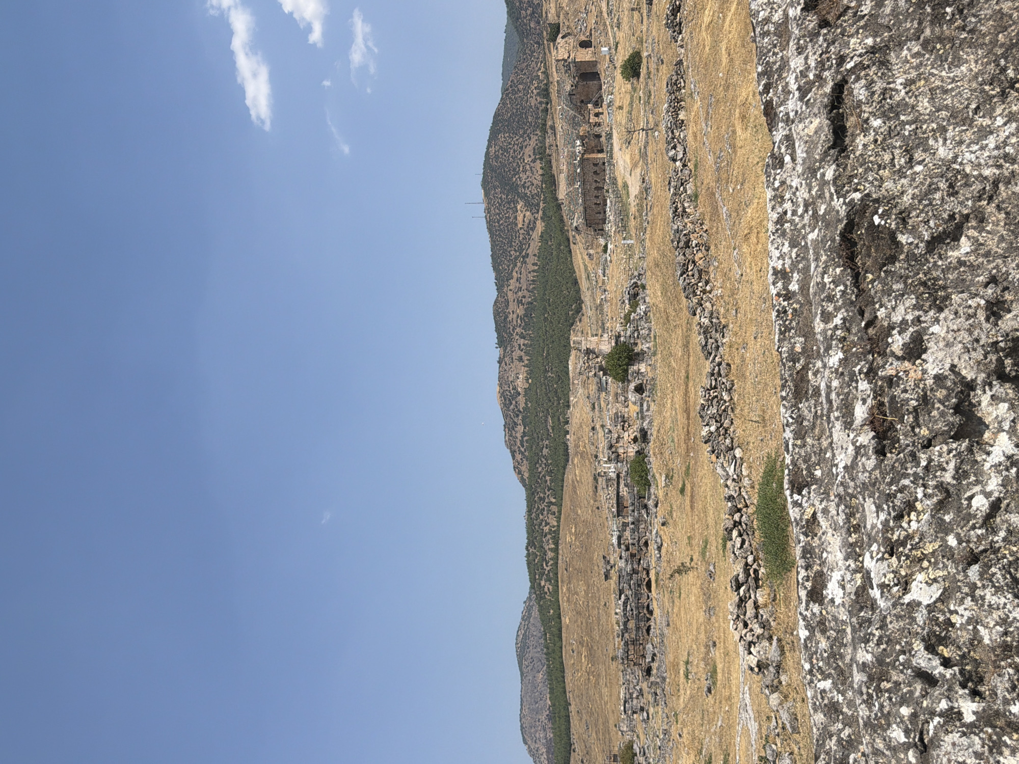 The image shows a wide shot of the Hierapolis ancient city in Pamukkale, Turkey, on a bright, clear day with a vast expanse of blue sky. In the foreground, a rough, textured grey stone structure dominates the bottom of the frame. Beyond this, a dry, golden-brown landscape stretches out, dotted with scattered grey stones and low, hardy green shrubs.
Further in the midground, ancient ruins are visible. These consist of stone structures, some appearing as arched foundations and others as weathered walls, hinting at past buildings. A low, meandering wall constructed from irregular stones runs horizontally across this area. In the background, rolling hills covered in dry grass and patches of darker green vegetation rise up, with more distant, larger hills forming the horizon line. A few scattered trees are present throughout the landscape. On the far right of the midground, a more substantial ancient building, possibly an amphitheater, with tiered seating and arched structures, is clearly discernible. A telecommunications tower is visible atop one of the distant hills. The overall scene depicts a historical archaeological site under a clear sky.