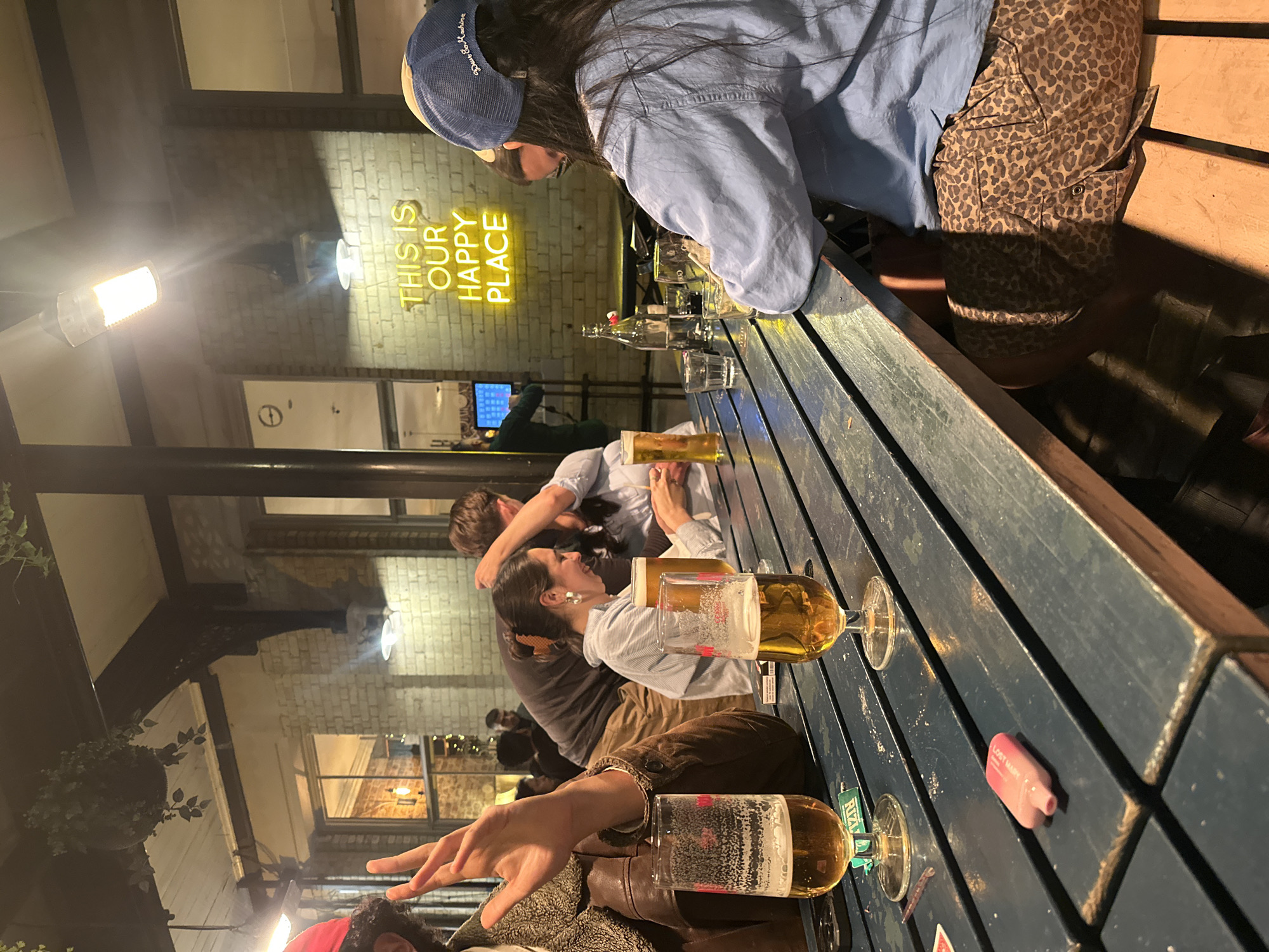 People are seated at a blue picnic table outside in the evening. On the brick wall behind them, neon lights spell out "THIS IS OUR HAPPY PLACE." Each person has a glass of beer, and there are bottles and smaller glasses on the table as well. One person is wearing a blue baseball cap and a denim jacket. The image seems to be taken in a casual, social setting at a pub or bar. Based on the style of brickwork and pub, the location could be in London, United Kingdom.
