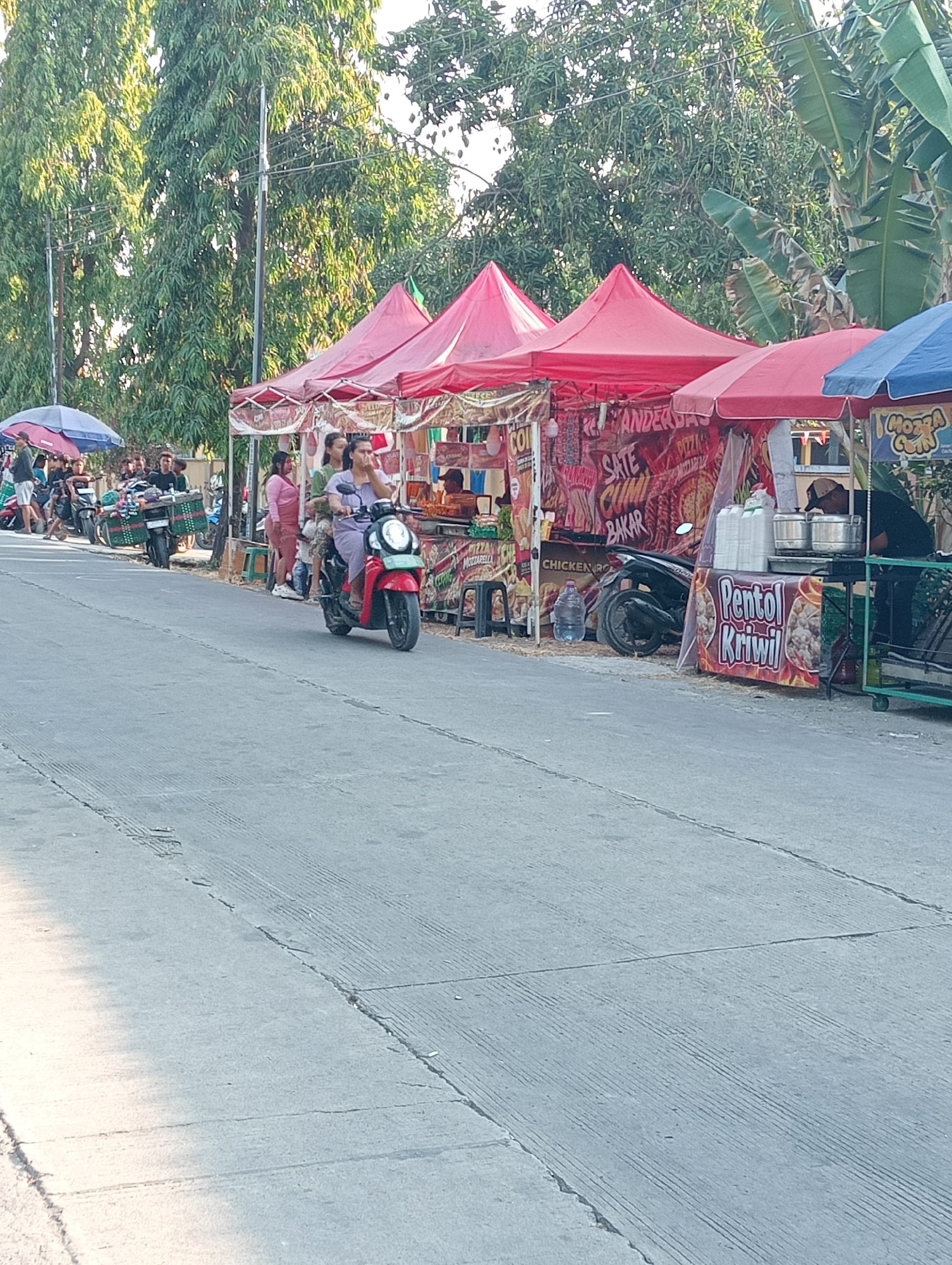 The image depicts a street scene in Tanjunganyar, Indonesia, lined with food stalls. Several red and blue awnings cover the stalls, displaying various food items and signage, including "Sate Cumi Bakar" (Grilled Squid Satay), "Chicken" and "Pentol Kriwil". People are seen gathered around the stalls and on motorcycles. One motorcycle with two passengers is in the foreground, moving away from the camera. Another motorcycle is parked next to a stall, with a vendor preparing food. The environment appears to be a bright, sunny day with lush green trees in the background. The road is paved, with distinct tire marks and cracks visible. The scene suggests a bustling market or street food area.