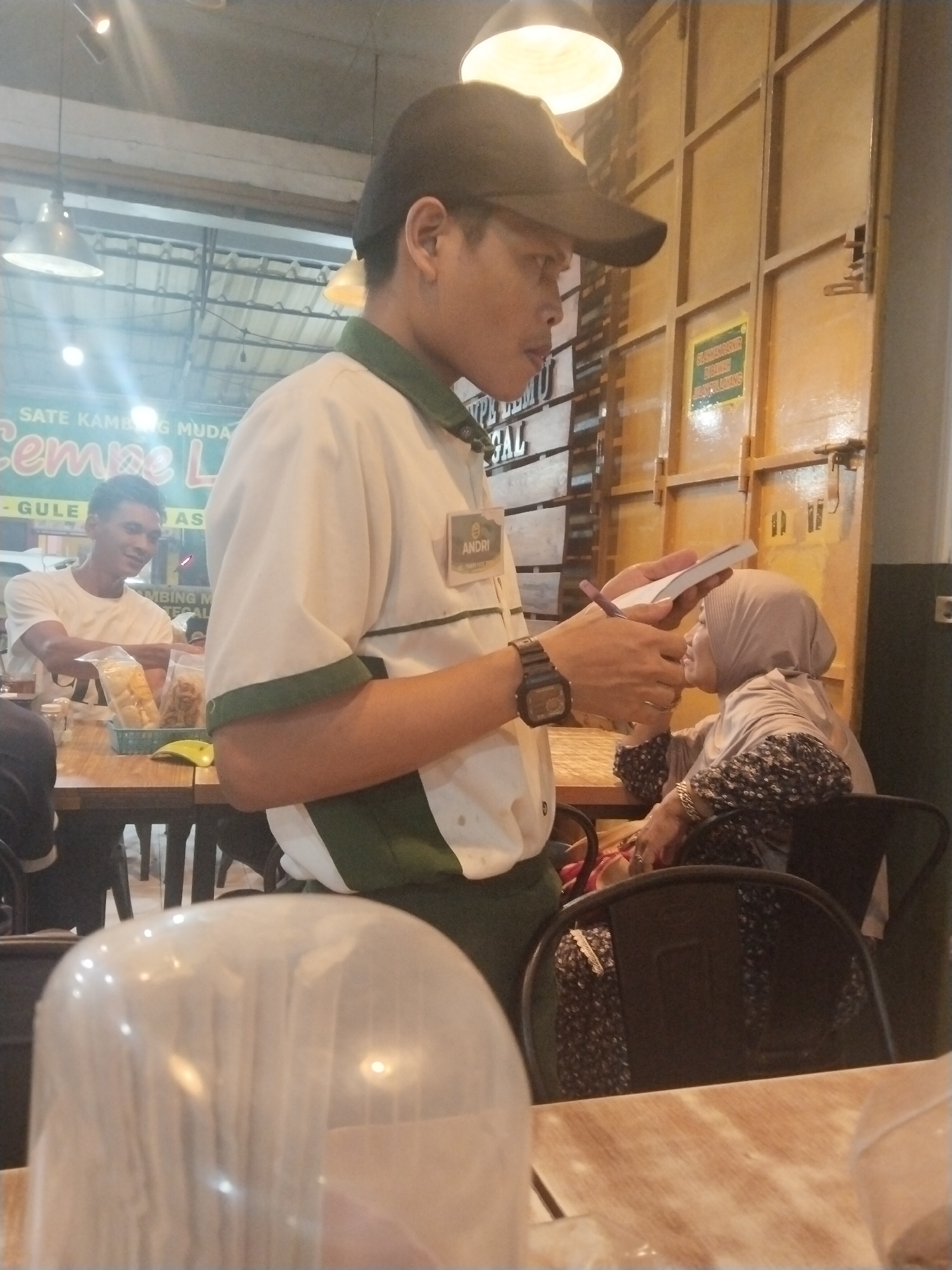An Indonesian man wearing a black baseball cap and a white polo shirt with green trim is standing and holding a small notebook and a pen, appearing to take an order or note information. He has a nametag on his shirt that reads "ANDRI". To his right, a woman wearing a light brown hijab and a patterned long-sleeved shirt is seated at a table, looking towards the man. The environment appears to be an indoor dining area of a restaurant, possibly in Adiwerna, Indonesia, with wooden tables and chairs, and overhead lighting. A clear plastic container is visible in the foreground.