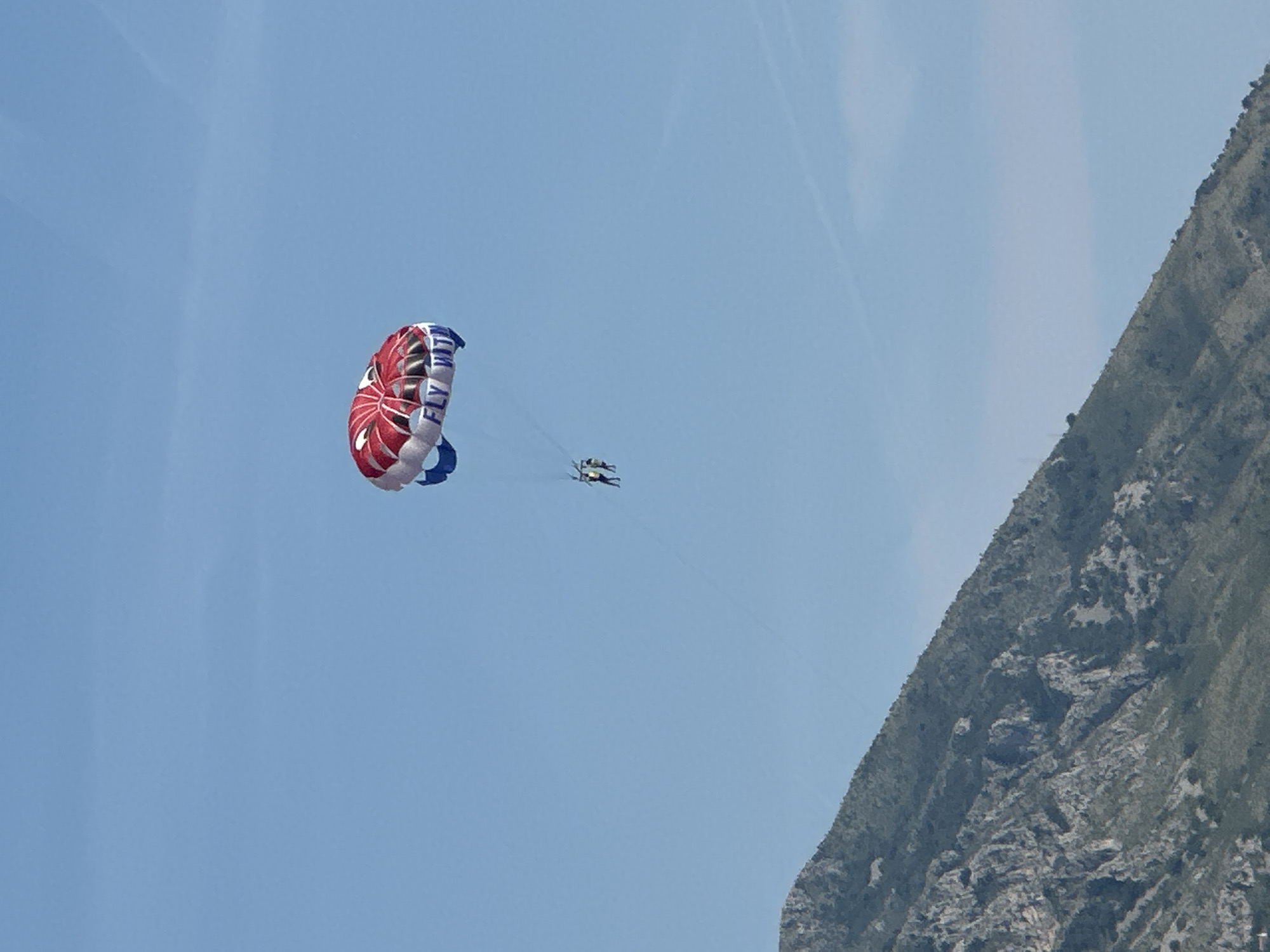 In the city of Boreti, Montenegro, two individuals are parasailing against a clear blue sky. They are attached to a large parachute-like canopy, which is predominantly red and white with blue trim. The canopy has the words "FLY WITH US" written on it in blue letters. Below the canopy, the two people are suspended in the air, appearing to be enjoying the activity. In the foreground, a portion of a steep, rocky mountain covered in sparse vegetation slopes upwards from the bottom left corner of the image. The mountain's texture is rough, with visible rock formations and shadows indicating uneven terrain. The overall scene suggests a daytime activity in a scenic coastal or mountainous region.