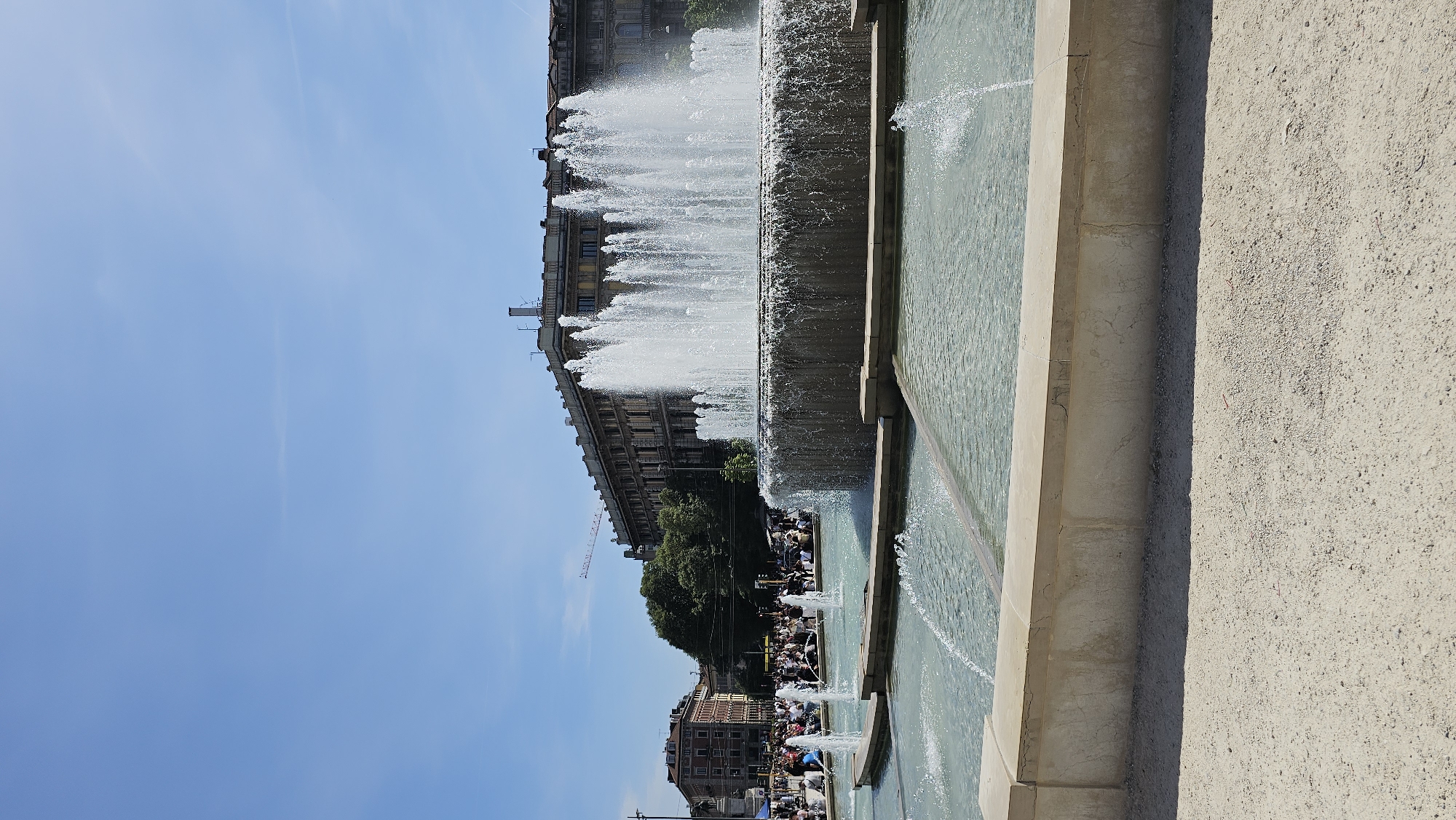 The image shows a tiered fountain in Milan, Italy, likely at Piazza Castello. The fountain is the primary subject, with water cascading down its levels. A crowd of people can be seen gathered around the fountain's base. Behind the fountain, buildings are visible, including a large, older structure. A construction crane is subtly visible in the background, suggesting urban development. The sky is clear and blue. A large concrete structure or building is in the right foreground.