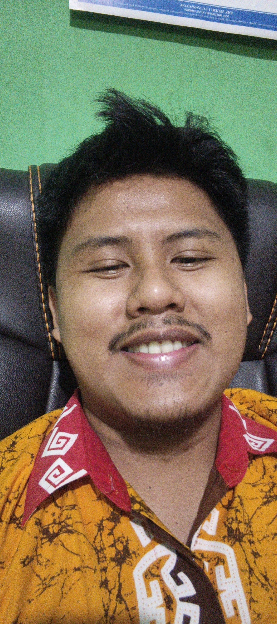 A close-up selfie of a man in Sukabumi, Indonesia, shows him seated in a black leather chair against a bright green background. The man has dark hair, a mustache, and is wearing a yellow shirt with a red collar adorned with white geometric patterns. He is looking directly at the camera with a slight smile, his eyes partially closed. The lighting is somewhat harsh, creating visible skin texture and highlights. A portion of a white sign with text in a foreign language is visible at the top of the frame.