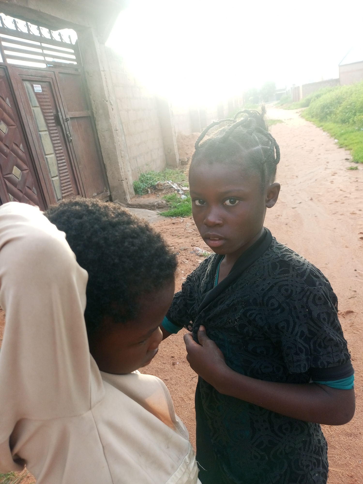 A young girl with braided hair stands on a dirt road in Tempure, Nigeria, looking towards the camera. She is wearing a black lace top over a teal undershirt. To her left, another child, facing away from the camera, is wearing a light-colored garment. In the background, a brick wall and a large metal gate are visible, along with vegetation and a building in the distance. The sun is bright, casting a strong glare on the scene.