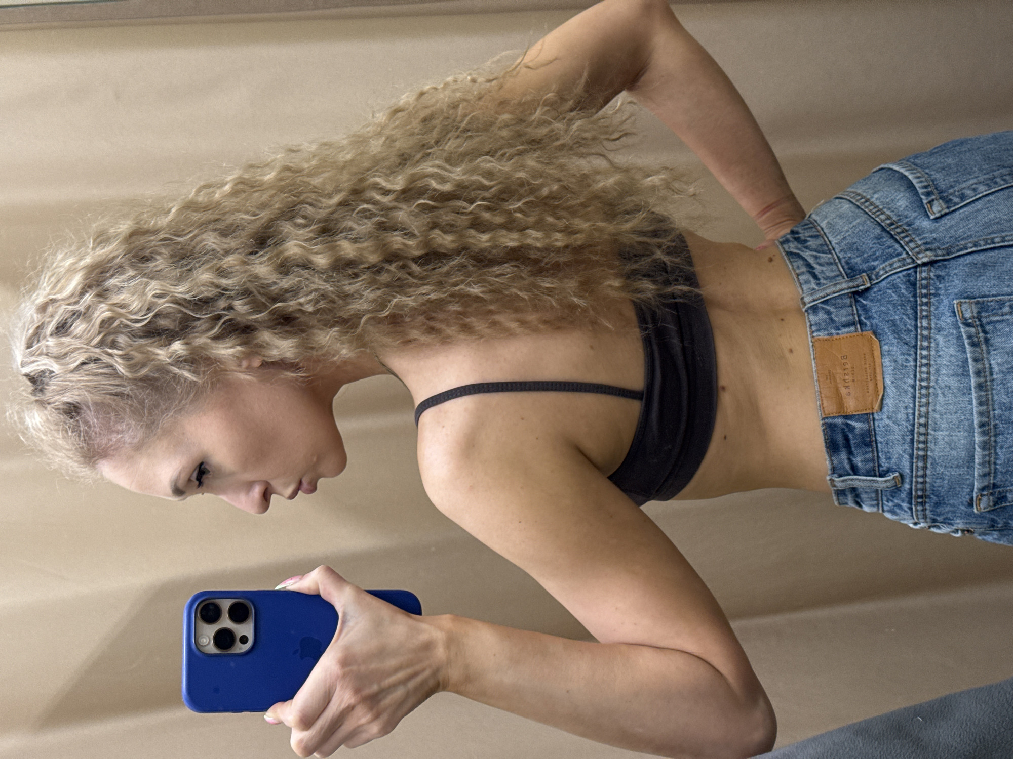 A young woman is reflected in a mirror while taking a selfie. She is wearing blue jeans and a black bra. Her long, curly hair is blonde. The woman is holding a blue smartphone in her right hand. The background is a neutral color. The image does not contain any information to indicate the location is in Perm, Russia.