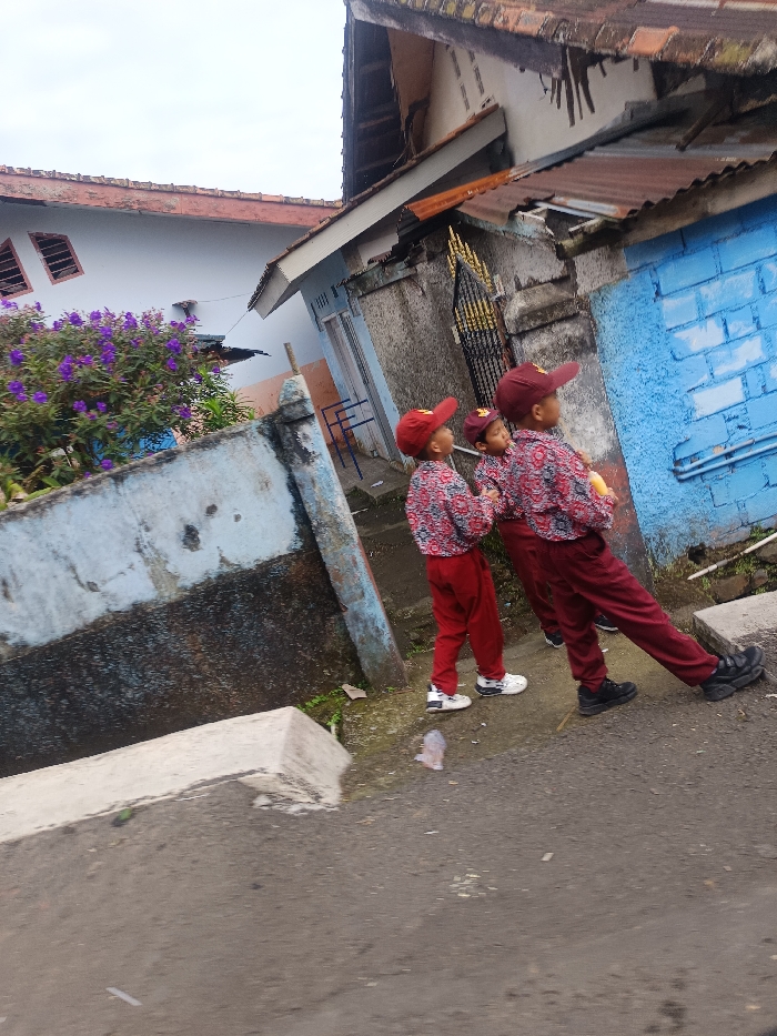 Three children in red uniforms and matching caps walk along a paved street. They are next to a low, weathered wall with dark stains and some greenery. Behind them are houses, one white with purple flowering bushes, and another with a blue wall and a rusty metal roof. The sky overhead is overcast.