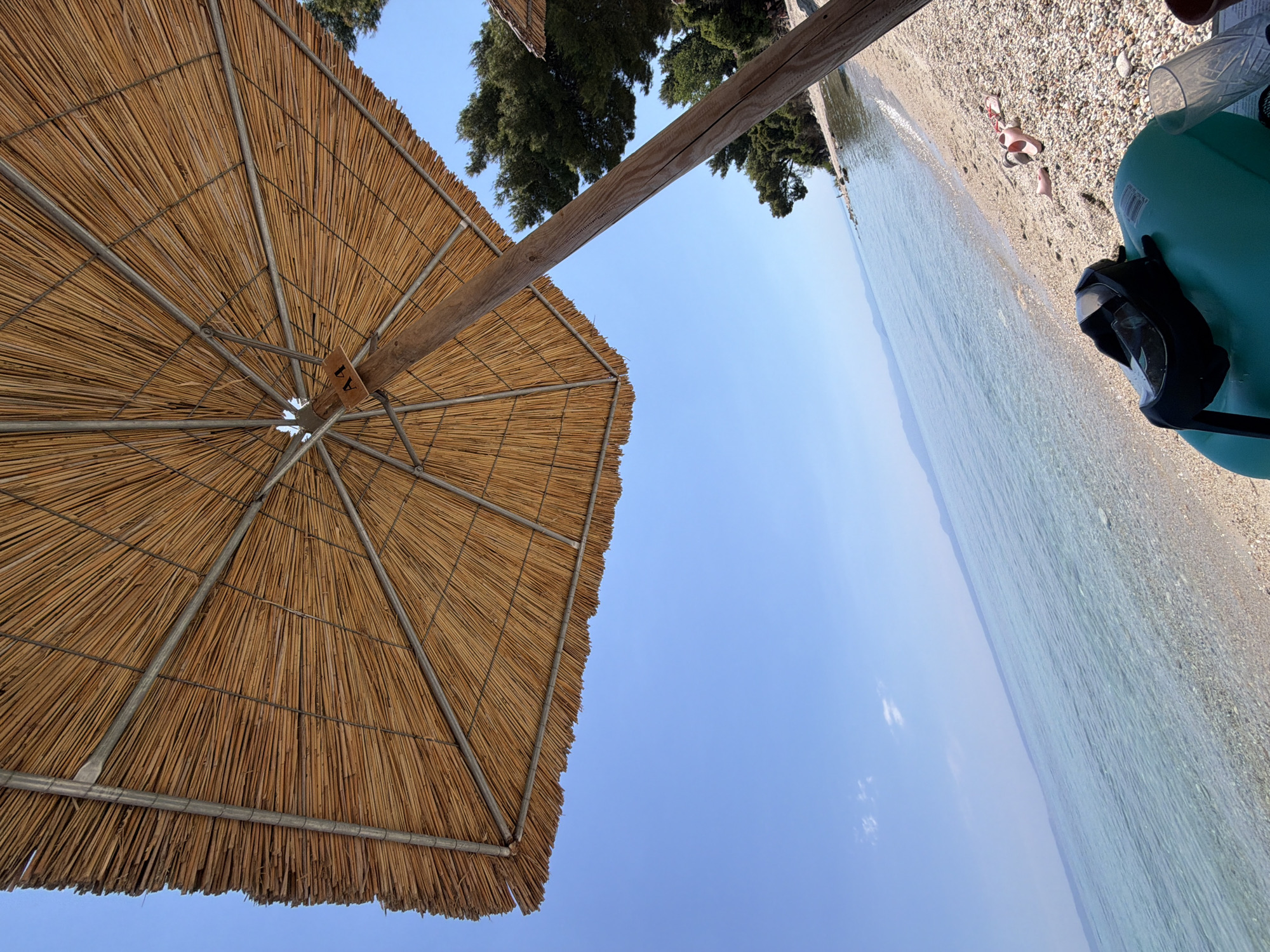 The image depicts a beach scene in Kassandra Municipal Unit, Greece, under a clear blue sky. The foreground is dominated by the underside of a large, thatched umbrella, supported by a wooden pole. The beach itself is composed of small pebbles and sand, with the calm, clear turquoise water of the sea gently lapping at the shore.
In the middle ground, on the beach, a pair of pink flip-flops lie near a snorkeling mask resting on a large, green cylindrical object. Further down the beach, two individuals are visible in the distance, seemingly relaxing. To the right, green pine trees line the edge of the beach, with another thatched umbrella visible further along the coastline. The horizon is marked by a faint, mountainous silhouette. The overall impression is one of a serene and idyllic beach setting.