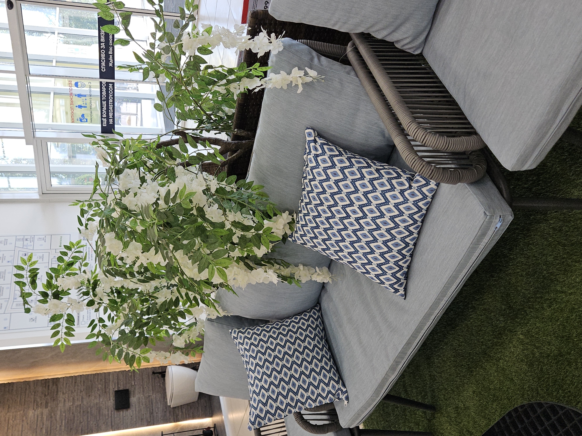 A modern seating area is depicted in what appears to be a commercial space in Ulyanovsk, Russia. The foreground features a grey upholstered sofa with two accent pillows, each adorned with a blue and white diamond pattern. The sofa has a woven grey armrest. The seating is placed on a vibrant green artificial turf rug. Behind the sofa, a large artificial plant with white flowers and green leaves spills over the furniture. In the background, a white toilet is visible on a grey tiled wall. Further in the background, automatic glass doors lead to an outdoor area. A sign with Russian text is visible on the glass doors, indicating a store or business. The overall impression is one of a stylish, indoor-outdoor display, possibly in a home improvement store or furniture showroom.