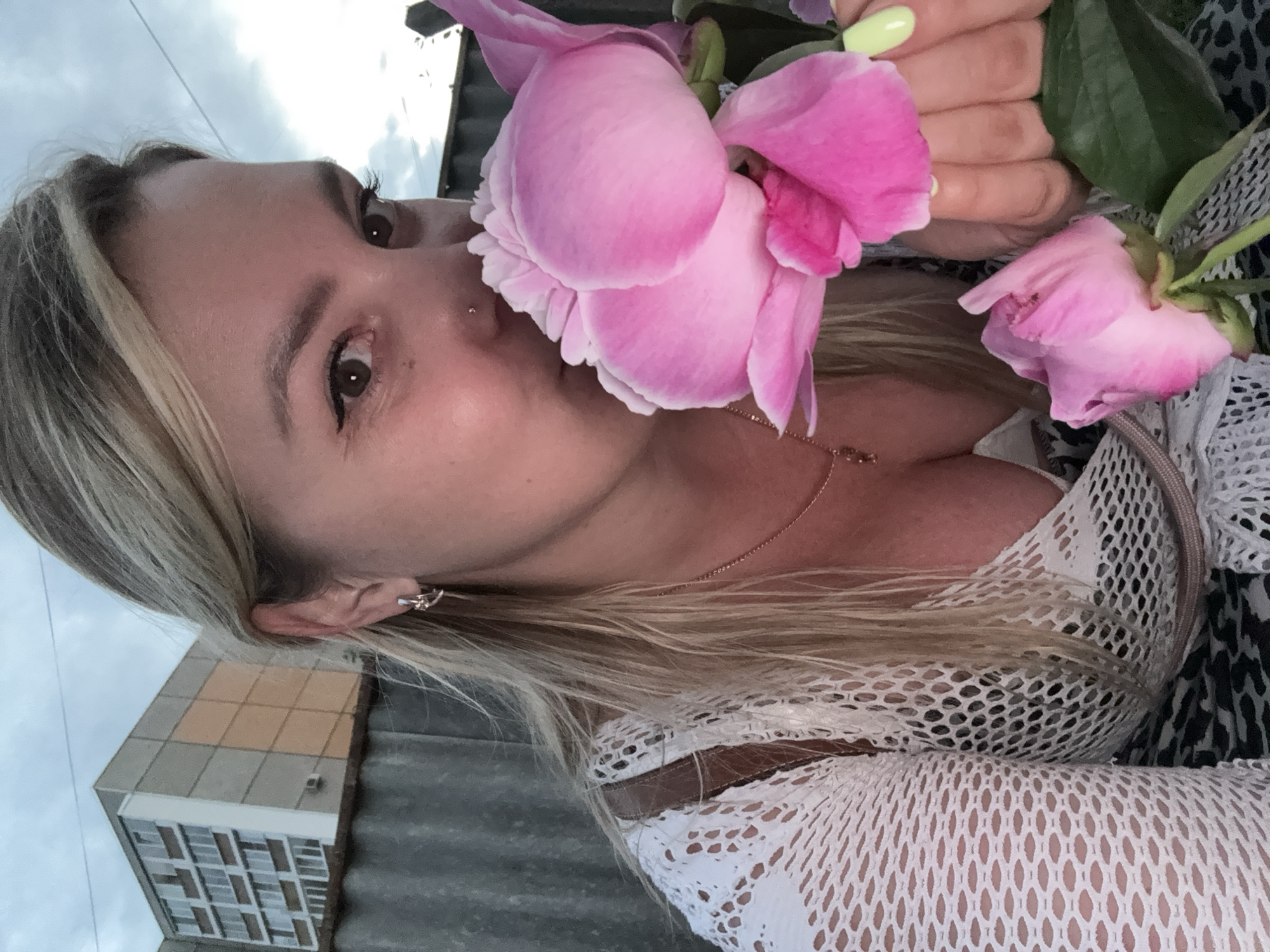 A woman with blonde hair and light skin is shown smelling pink peonies. She is wearing a white mesh top and a necklace. Her ear has a small earring, and she has a nose ring. In the background, there is a gray fence and a building with a gray and orange facade. The sky is cloudy and gray. The location is Chaikovsky, Russia.