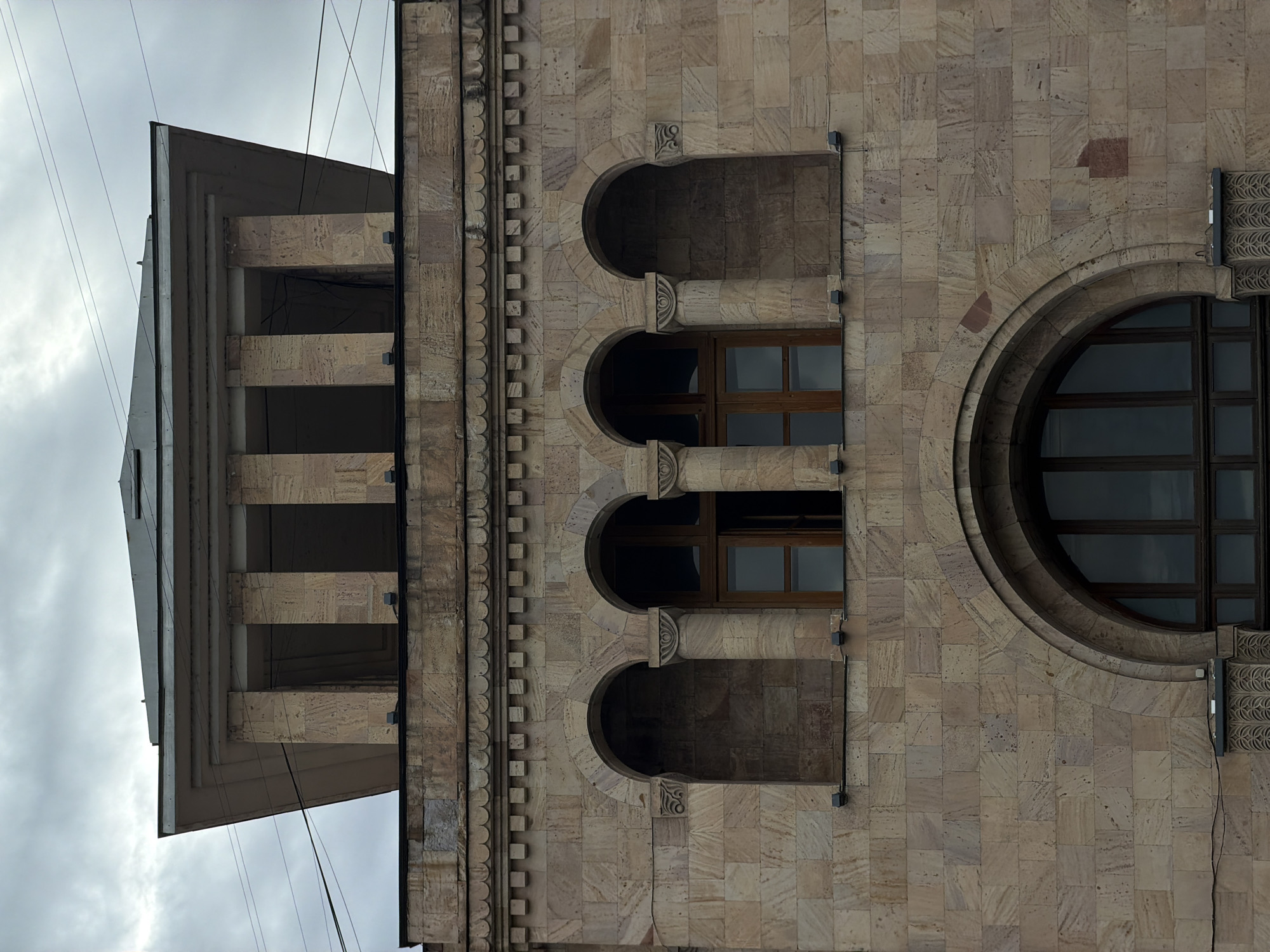 The image depicts a section of a stone building with distinctive architectural features, including a tower-like structure with horizontal divisions, a series of arched window openings, and a large circular window. The building's facade is composed of light-colored, textured stone blocks, with ornamental details along the edges. The sky above is overcast. The building is the National Academic Theatre of Opera and Ballet in Yerevan, Armenia.