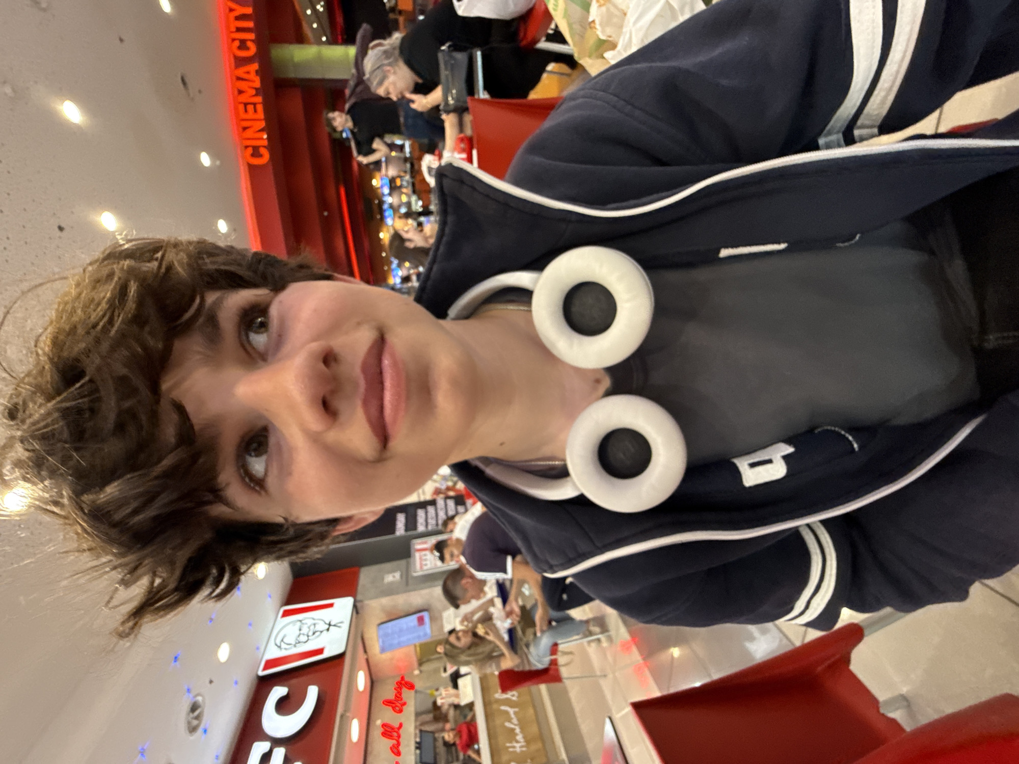 A young person with short, dark, curly hair is taking a selfie. They have light skin and are wearing a gray shirt, a navy blue jacket with white stripes, and white headphones around their neck.
Behind the person, there's a busy fast food restaurant. Visible signs indicate it's a KFC, with menus and promotional signage in the background. Red seating is present.
In the upper part of the image, "CINEMA CITY" is visible in red lettering, suggesting the KFC is located near or inside a cinema complex. Other people are seen dining in the background.