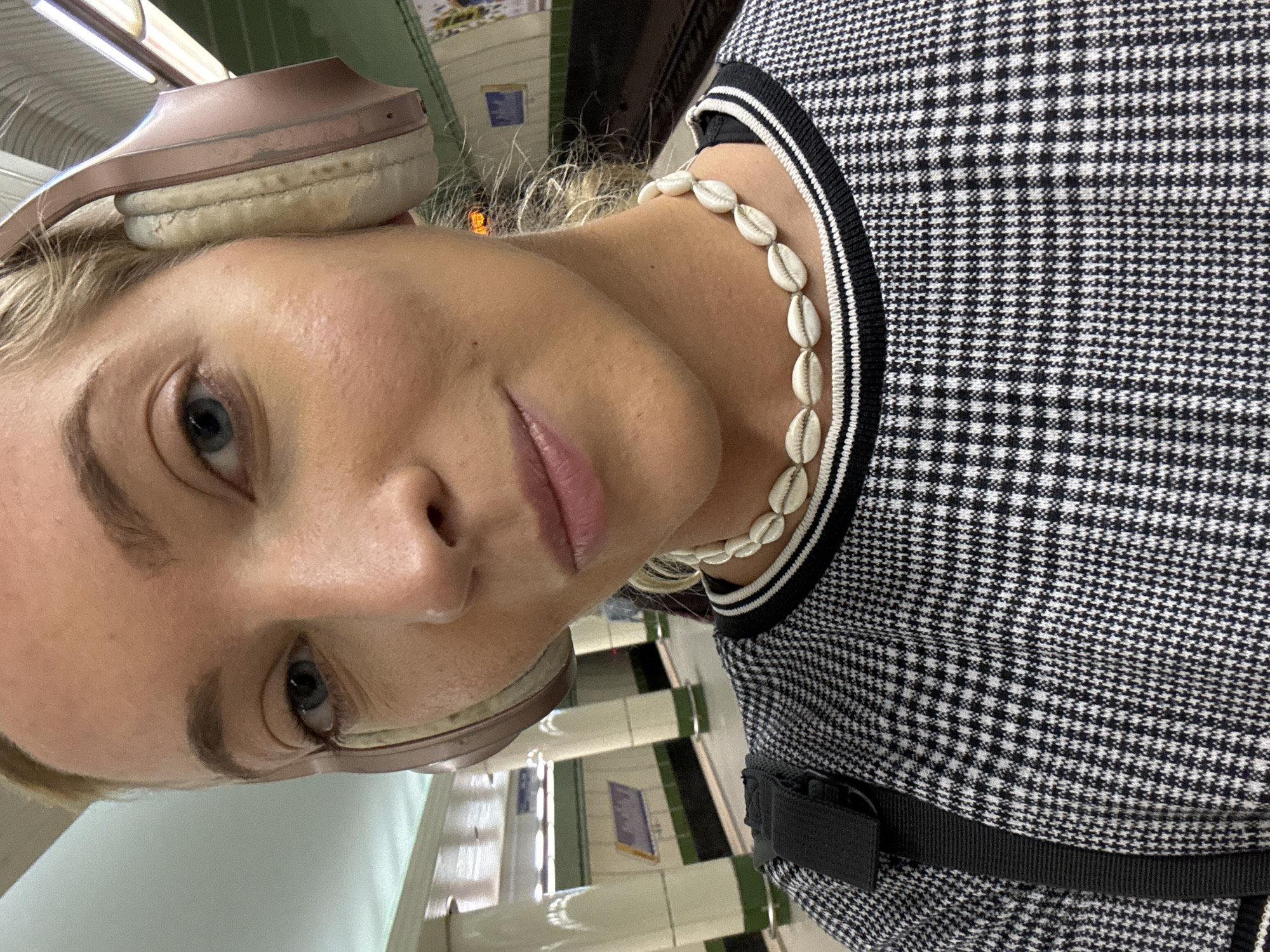 A selfie was taken by a female at a location in Minsk, Belarus. The selfie is of a fair-skinned female with light-blonde hair. She has headphones over her head and a necklace of white shells around her neck. She wears a black and white checkered pattern top. The background contains the interior of a Minsk subway/metro station, displaying white pillars, and tiled walls in white, green, and black. There is a sign present in the background.
