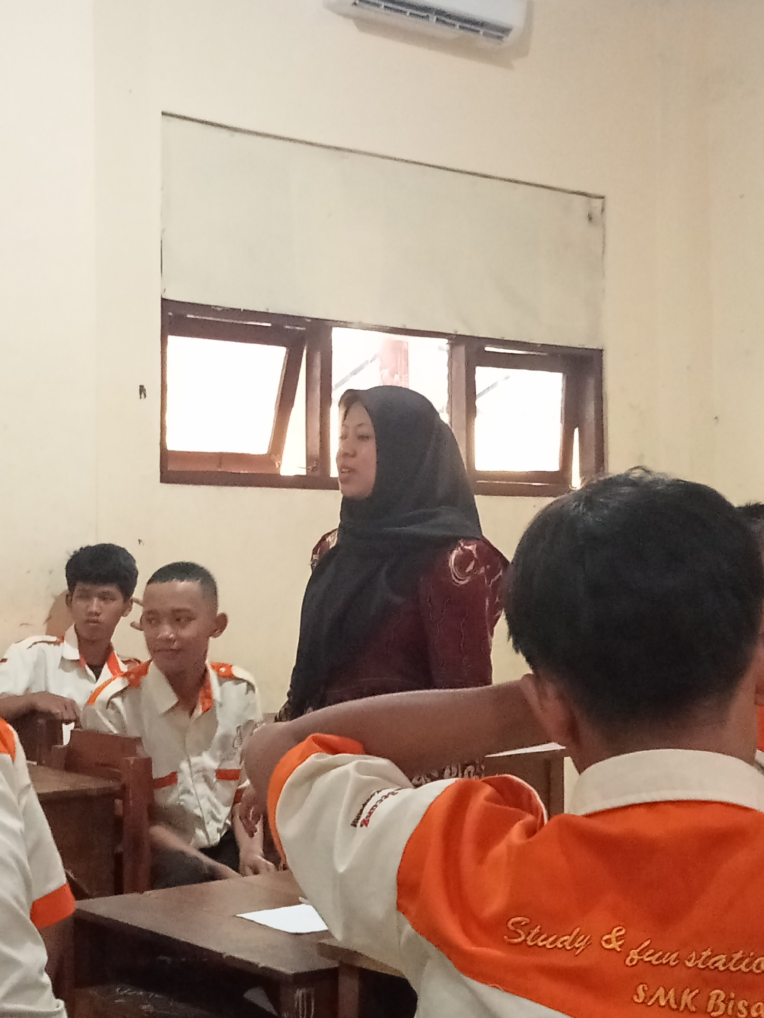 In a classroom in Secang, Indonesia, a young woman wearing a black hijab and a maroon patterned top stands facing a group of students seated at desks. She appears to be speaking or instructing. The students are wearing white shirts with orange trim, possibly a school uniform. Some of the students are looking towards the woman, while others are looking down or away. The classroom has windows with wooden frames, and an air conditioning unit is visible on the wall. The overall scene suggests an educational setting, likely a school.