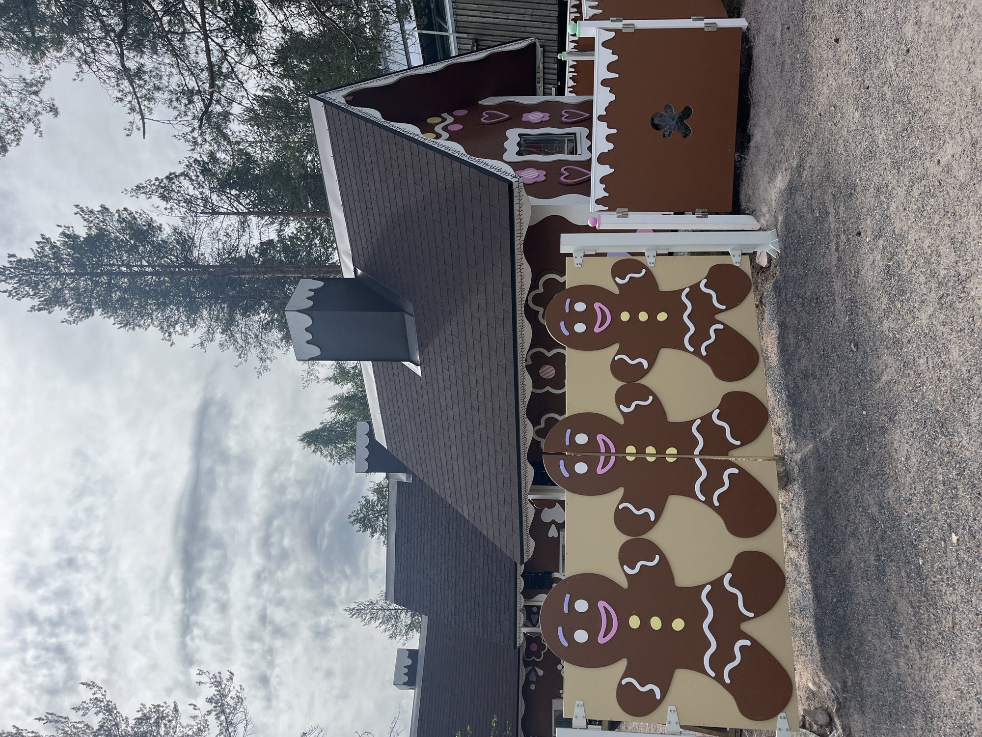 The image shows a facade of a gingerbread house-themed building located in Rovaniemi, Finland. The building has a dark brown roof, white trim, and decorative elements resembling candy. In the foreground, there is a gate featuring three large, cartoonish gingerbread man cutouts. The gate is tan and the gingerbread men are brown with white, yellow, and pink details for their faces and clothing. To the right of the gate, a section of brown fence has a cutout in the shape of a gingerbread man. The ground is covered in gravel. Tall pine trees are visible in the background, and the sky is overcast. The scene appears to be an outdoor attraction or themed area.