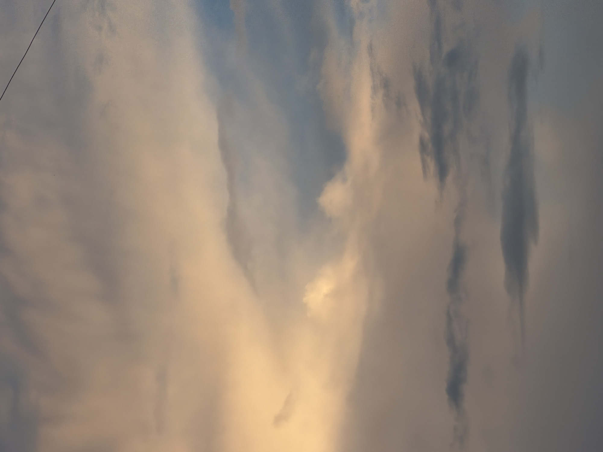 The image shows a cloudy sky with warm, golden light illuminating some of the clouds from the left, suggesting it may be sunrise or sunset. There are darker, grey clouds scattered throughout the frame, with a thin wire visible in the upper right corner. The lower part of the image is darker, with more defined, low-lying grey clouds. There are no people or specific landmarks visible to indicate a location in Khimki, Russia.