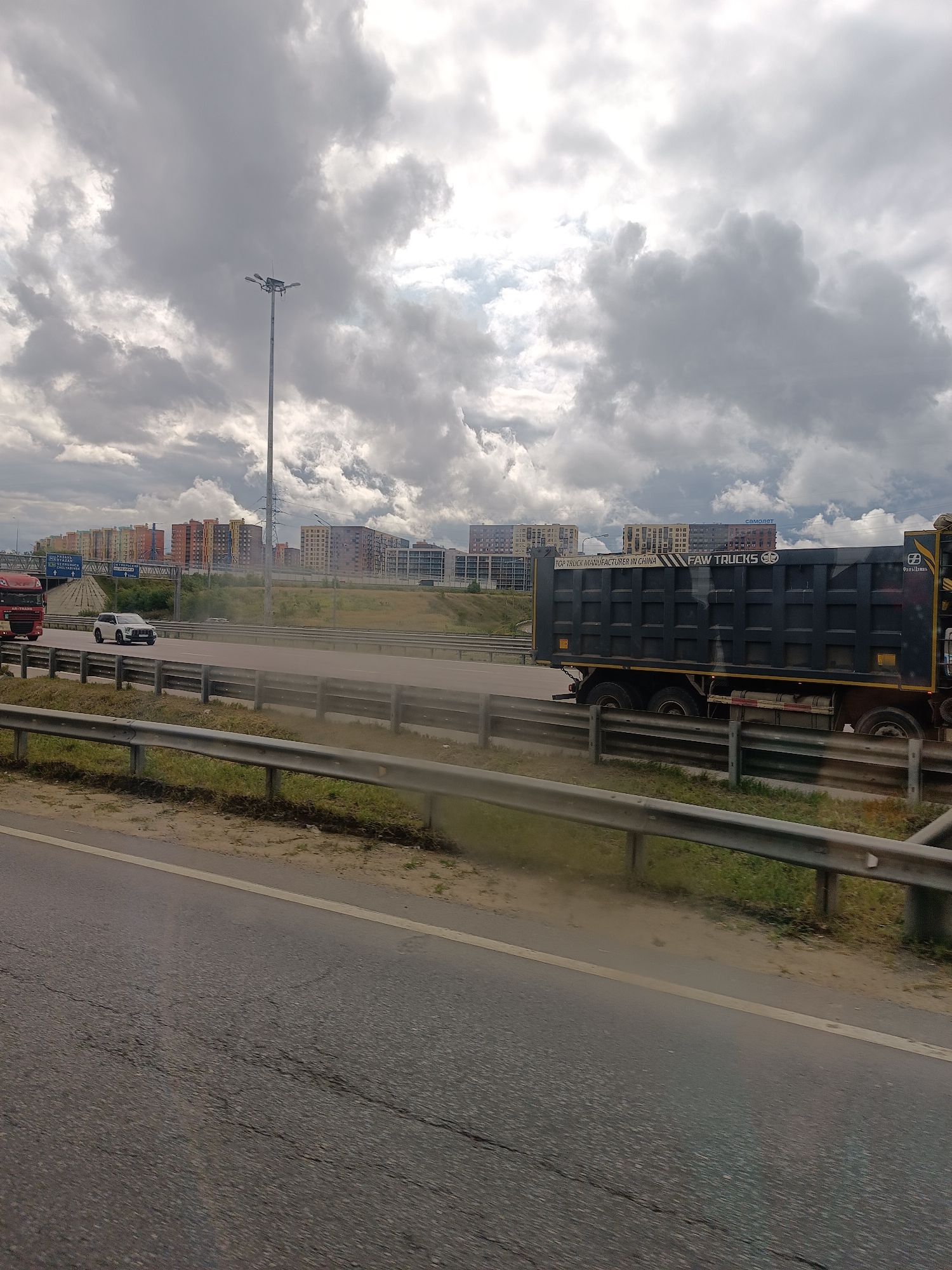 The image depicts a highway scene in Zhilino-1, Russia. In the foreground, a cracked asphalt road is visible, bordered by a metal guardrail and dry grass. On the highway, a white SUV is traveling to the left, and a red truck is to its left. Further ahead, a dark grey dump truck with "FAW TRUCKS" branding is also moving. In the background, a cluster of multi-story residential buildings with colorful facades is visible under a cloudy sky. A tall lamppost stands near the buildings, and power lines are also present. The overall environment suggests an urban or suburban setting with ongoing construction or development.