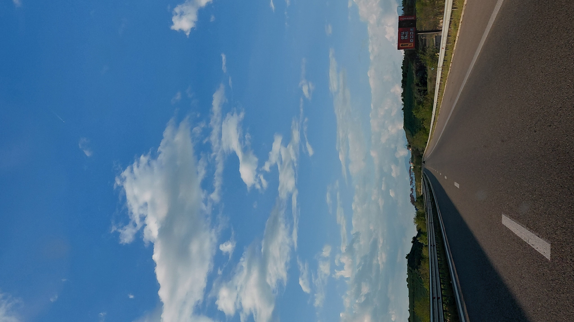 The image depicts an open highway on a clear, sunny day, captured from a low perspective, possibly from a moving vehicle. The road, made of asphalt, extends from the foreground into the distance, curving gently to the right. White dashed lines mark the lanes, and a solid white line runs along the right shoulder, bordered by a metal guardrail.
To the right of the road, the landscape features lush green hills covered with trees and vegetation. Further in the distance, what appears to be a body of water, possibly a river or lake, is visible, reflecting the bright sky. A large red and white billboard stands prominently on the right side of the highway.
The sky dominates the left side of the image, a vibrant blue adorned with numerous scattered white cumulus clouds of varying sizes and shapes. A faint white contrail from an airplane is visible in the upper left portion of the sky.
The weather is bright and clear, indicating daytime, likely late morning or early afternoon given the high position of the sun and the shadows cast.
Visible text on the billboard includes "TOSKOGO" at the top, "GRILL GRILL" written twice below it, and "X CC P" below "GRILL". An arrow on the billboard points to the left.