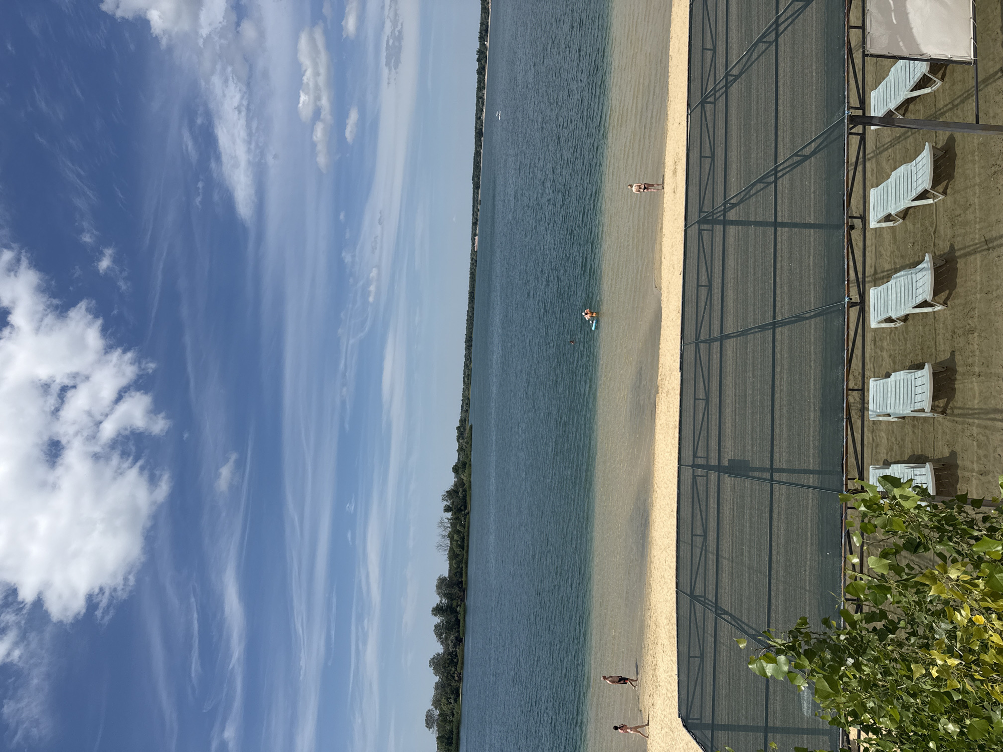 The media file shows a beach scene on a sunny day, likely in the afternoon given the shadows. The sky is a bright blue with wispy white clouds. The water of a lake or sea is calm and a deep blue-green color, meeting a sandy beach. Along the edge of the beach, a fence-like structure with a dark mesh is visible, behind which are several white lounge chairs, suggesting a recreational area.
There are a few people visible on the beach. In the shallow water, one person appears to be wading or swimming. Further up the beach, closer to the water's edge, another person is standing. Two more individuals are seen walking on the sand towards the right of the frame. The general atmosphere seems relaxed and leisurely, typical of a beach day. The presence of trees in the distance along the shoreline indicates a natural, possibly wooded, environment surrounding the beach. The location is identified as Yelyzavetivka, Ukraine. There is no visible text in the image.
