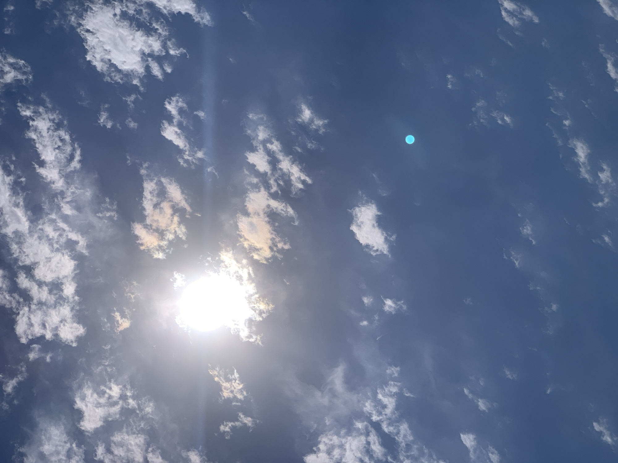 The image shows a bright sun partially obscured by clouds, casting light across a blue sky. Scattered white and light orange clouds are visible throughout the frame. In the lower right quadrant, a distinct bright blue, circular object is present, possibly a lens flare or a reflection. The scene appears to be an outdoor, daytime sky. There are no people, subjects, objects, or actions depicted other than the natural phenomena of the sky. There is no visible indication of the city of Khazar Raion, Azerbaijan.