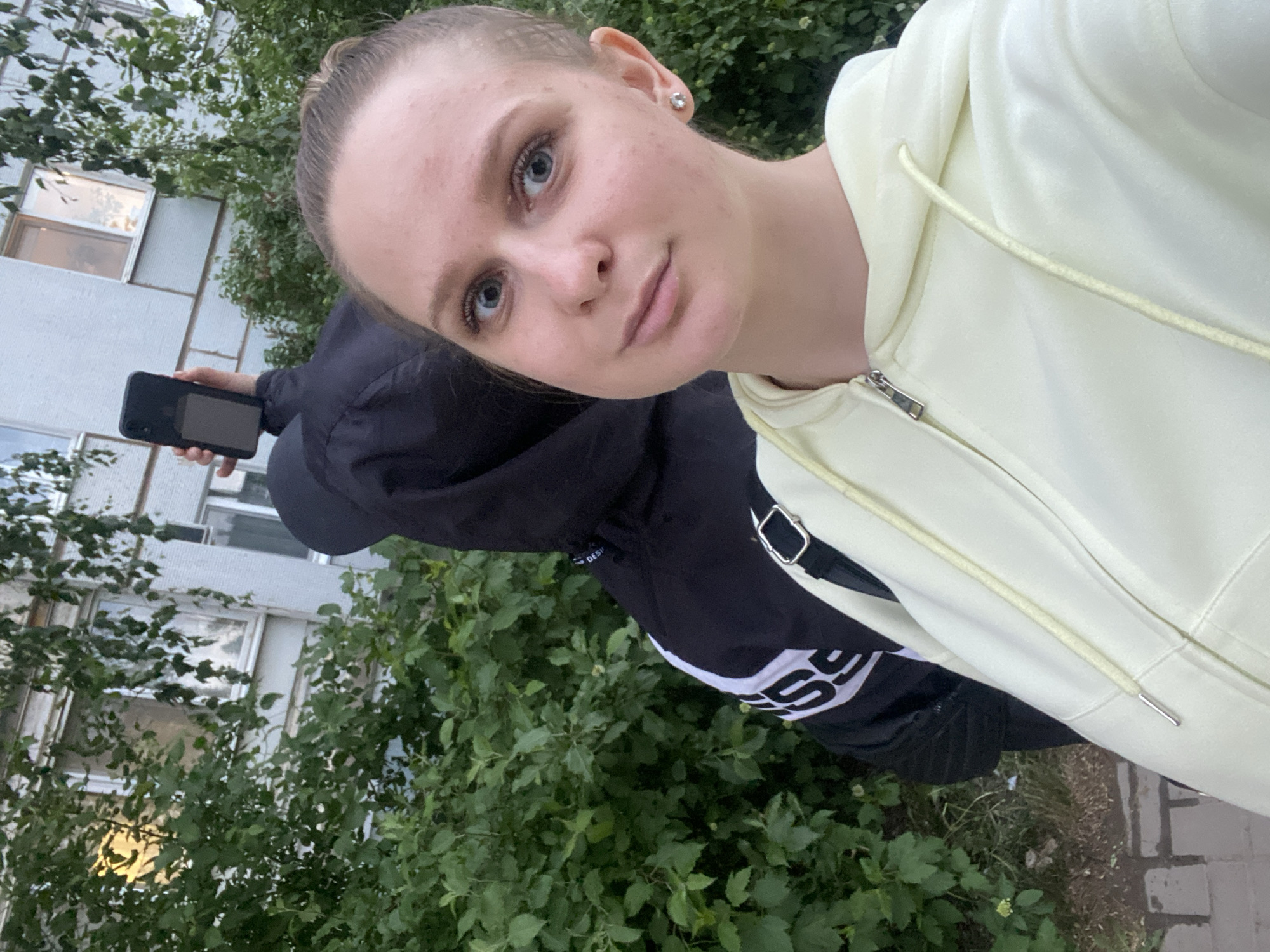 A young, light-skinned woman with blonde hair pulled back is taking a selfie. She has blue eyes and is wearing a light yellow zip-up hoodie. To her left and slightly behind her, a person wearing a black hooded jacket holds a black smartphone. They are standing in front of green foliage. In the background, there's a multi-story building with windows. The scene appears to be outdoors in the city of Chaikovsky, Russia.
