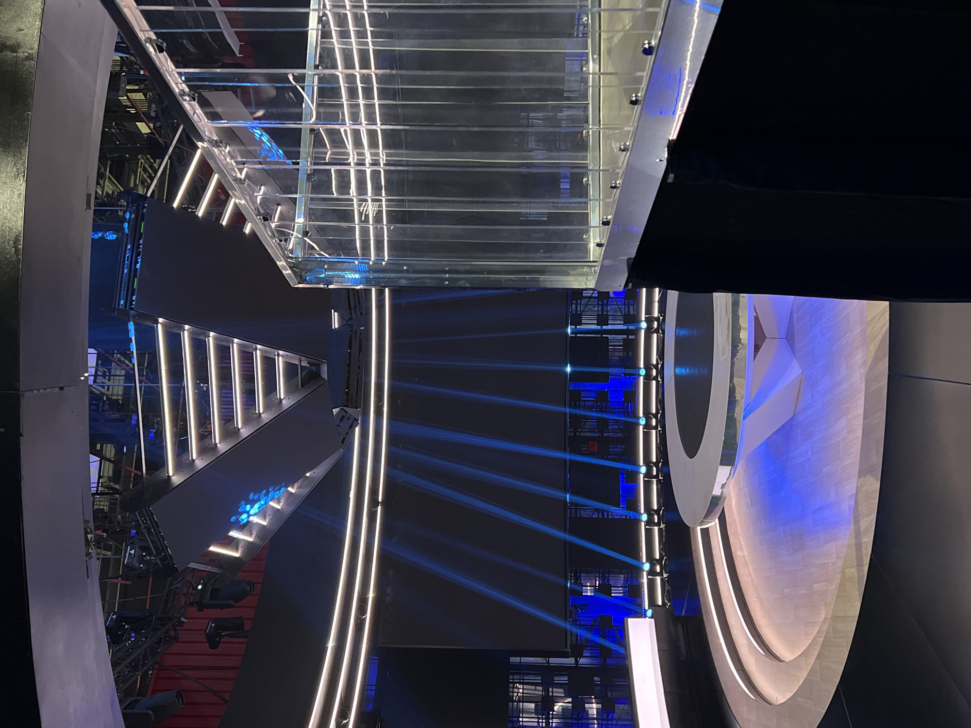 The image displays a multi-level, intricately designed stage setup within a large indoor venue, likely a studio or performance hall, in Khimki, Russia. No people are visible.
The scene is illuminated primarily by artificial blue and white light, creating a dynamic and modern atmosphere. A prominent feature is a transparent or reflective upper walkway, possibly a bridge or set piece, characterized by a grid pattern and subtle reflections of light. Below this, various platforms and angled structures are integrated with vertical strips of white light, outlining the stage architecture.
Numerous beams of blue light cut across the dark background, originating from unseen light fixtures and enhancing the visual effects. On the right side of the frame, a large, curved, and layered backdrop or scenic element is visible, illuminated with a soft blue glow. In the background, scaffolding and other technical equipment like speakers and lights indicate a complex production environment.
The overall impression is that of a professional stage, meticulously prepared for a large-scale event such as a concert, television show, or awards ceremony. The lighting and structural design suggest a focus on visual spectacle and a high-tech aesthetic. The photograph is taken from an elevated perspective, looking down onto the stage area. No visible text is present in the image.