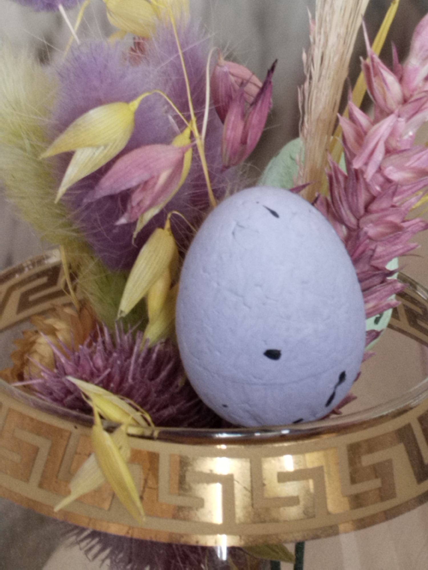 The image displays a close-up of a decorative Easter egg, approximately life-sized, nestled within a floral arrangement. The egg is a pale lavender color with several small, irregularly shaped black spots scattered across its surface. It is positioned in the foreground, slightly off-center to the left.
Behind the egg, a collection of dried and possibly dyed botanicals creates a textured backdrop. Dominant colors include soft purples, pale yellows, and dusty pinks, with some hints of light green and beige. These elements appear to be arranged in a bouquet or wreath formation. Specifically, there are feathery, plume-like elements in purple, elongated seed pods in yellow and pink, and spikey, thistle-like blooms in purple and pink. A strand of dry, straw-like material is also visible.
The arrangement rests within a decorative bowl or stand. The visible portion of this container is a band with a metallic gold finish, featuring a Greek key pattern in a darker, muted gold or bronze. The bowl appears to be made of glass, as evidenced by a slight sheen and reflection.
The lighting in the image is soft and diffused, suggesting an indoor setting. The background is out of focus, providing no specific environmental context beyond the immediate arrangement and its container. There are no people or animals present in the image, nor are there any discernible actions or interactions taking place. The scene is static and appears to be a still life composition.
The image does not contain any information that would allow for the identification of its location in Hrodna, Belarus.