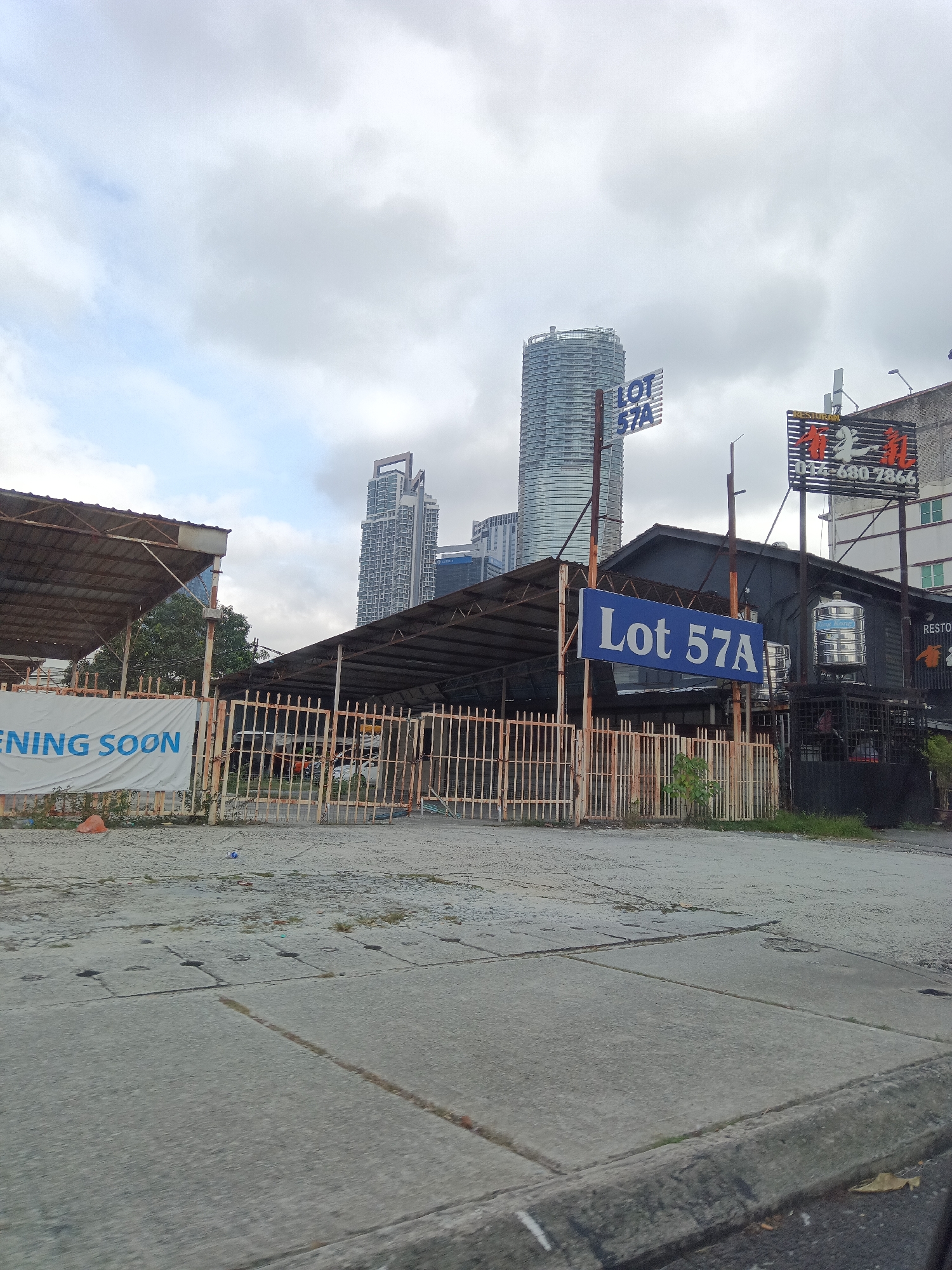 The image depicts an overcast day in Kuala Lumpur, Malaysia, with a focus on a commercial lot and surrounding structures. In the foreground, a concrete pavement shows signs of wear and tear with visible cracks and drainage openings. A low concrete curb separates the pavement from a blurred area on the right, suggesting it was taken from a moving vehicle.
Dominating the mid-ground is a fenced-off area with a rustic, weathered appearance. A long, covered structure with a corrugated metal roof, supported by metal poles, spans across the lot. Adjacent to this is a smaller, darker building with a similar roof structure. A large white banner with blue lettering that reads "ENING SOON" is displayed on the fence to the left, indicating an upcoming opening.
Several signs are visible in the image. A blue sign with white lettering reading "Lot 57A" is prominently placed on a pole in front of the darker building. Another smaller blue sign with the same text, "LOT 57A", is situated higher up on a pole to the left. To the right of the dark building, a taller sign with Chinese characters and the phone number "017-680 7866" is visible, with the word "RESTORAN" in smaller text above it. A stainless steel water tank is mounted on the side of the dark building.
In the background, the distinctive architecture of Kuala Lumpur's skyscrapers pierces the cloudy sky. A tall, cylindrical building with a unique, layered facade stands out, and another modern skyscraper with a more angular design is also visible. The overall impression is a developing or existing commercial area in an urban setting, marked by both the ongoing development (indicated by the "ENING SOON" banner) and established businesses.