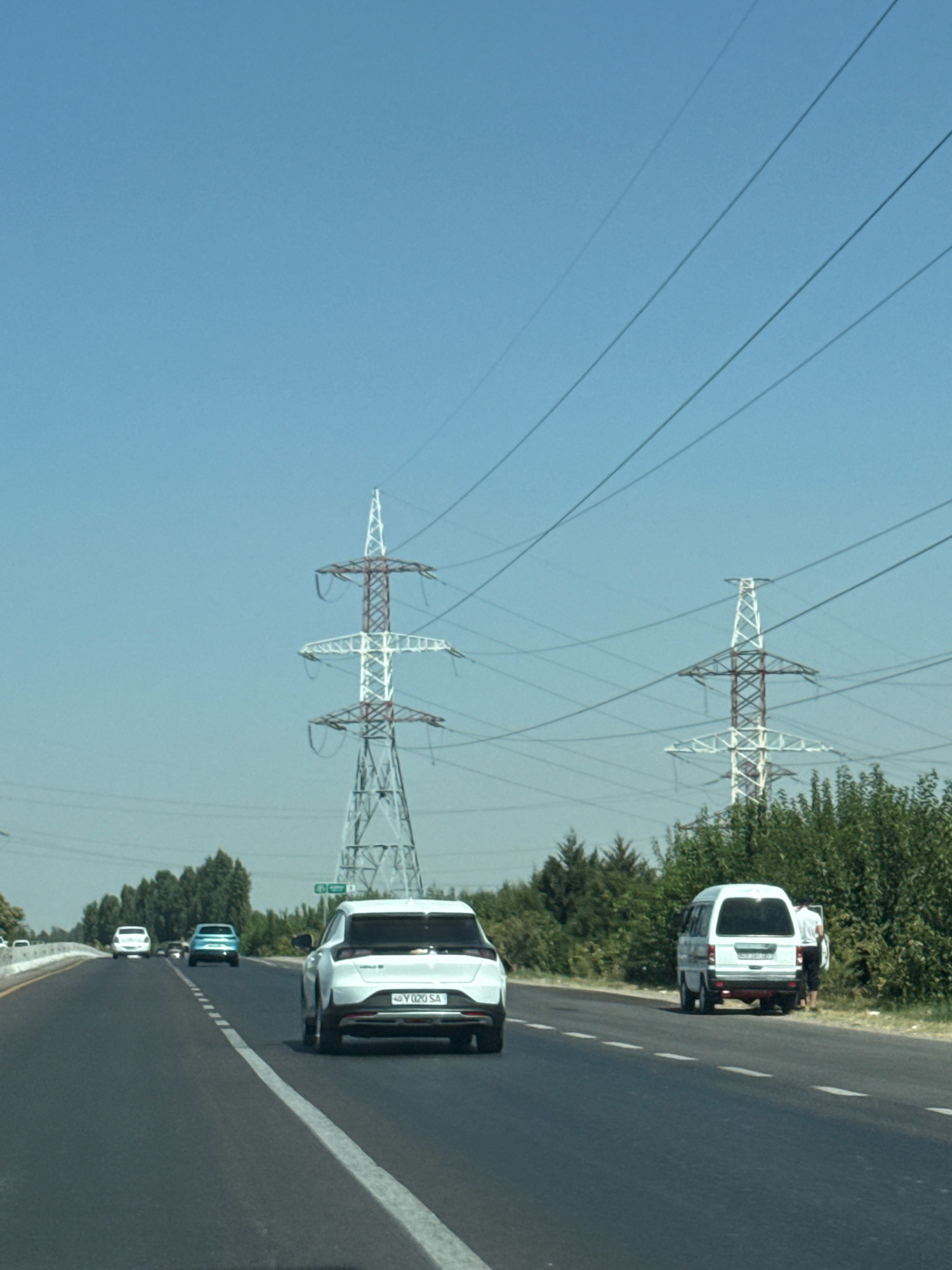 A daytime, outdoor, eye-level, horizontal, medium shot captures a highway scene in Istiqlol, Uzbekistan, dominated by a clear blue sky and a series of power lines supported by tall, lattice-like transmission towers. The road, paved with asphalt, features white lane markings and divides into multiple lanes. Several vehicles are visible on the road. In the foreground, a white SUV is driving away from the camera. Further down the road, a white minivan is parked on the shoulder, with two individuals standing near it. A light blue sedan is in motion ahead of the white SUV. In the middle ground and background, lush green trees line the sides of the road, contrasting with the skeletal structures of the power towers. The overall impression is of a functional transportation artery set against a backdrop of both modern infrastructure and natural greenery under a bright, cloudless sky.