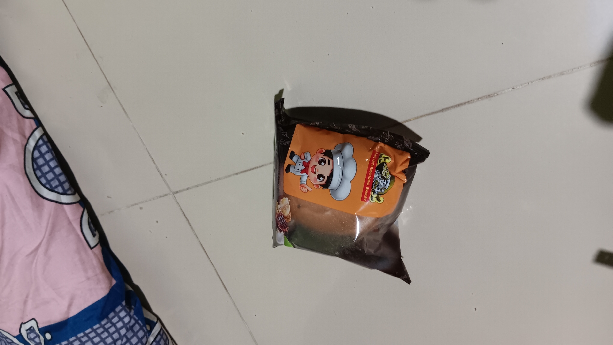 A package of bread sits on a tiled floor. The package is mostly brown and orange and features a cartoon image of a chef. The bread inside appears to be a light brown color. To the right of the bread package is a pink and blue patterned blanket. The scene is lit by overhead lighting, casting shadows on the floor. The image appears to have been taken in a residential setting in Sonobekel, Indonesia.