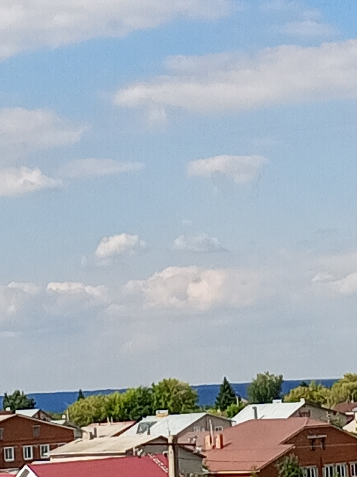 The image captures a residential area in Chistopol, Russia, under a partially cloudy sky. The foreground features rooftops of several houses, predominantly with red and white coverings. The houses appear to be of various architectural styles, with some displaying brick exteriors and others lighter-colored walls. Chimneys are visible on some of the roofs. In the background, a line of green trees forms a natural boundary, behind which a body of water, likely a river or lake, is visible. The sky above is a pale blue with scattered white clouds of varying shapes and sizes. No people are present in the image.