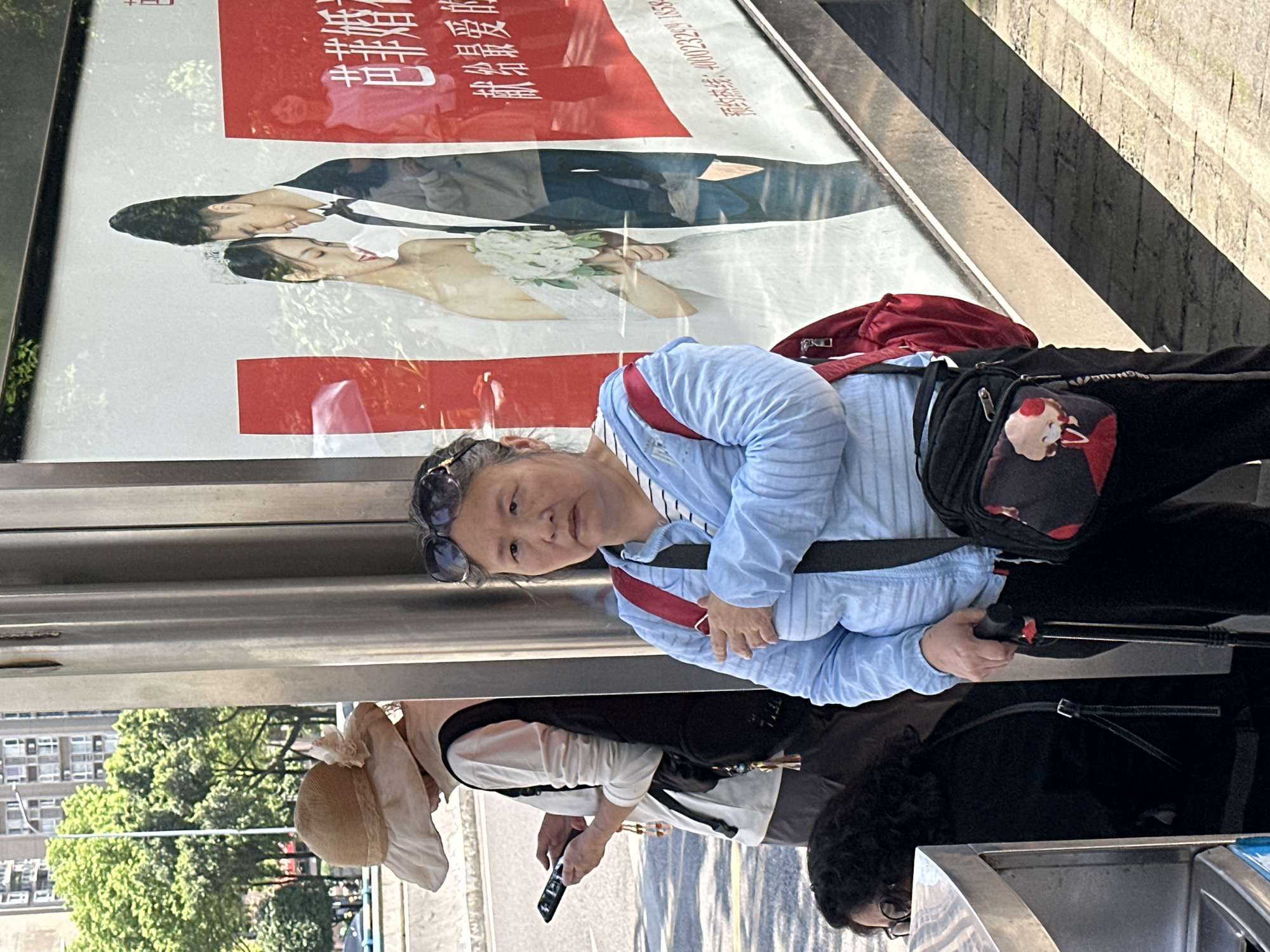 The image, taken in what appears to be Shapingba District, China, shows two women standing at what seems to be a bus stop or similar public transit shelter. One woman in the foreground is wearing a light blue long-sleeved shirt, dark pants, and a dark bag with a cartoon character on it. She has sunglasses on top of her head and appears to be looking directly at the camera. She is holding the handle of a wheeled suitcase. Behind her, another woman is visible wearing a straw hat and a white top with a dark vest. She is holding a mobile phone. The backdrop of the scene is a bus shelter advertisement, featuring a wedding photo with Chinese characters. The setting includes buildings and trees visible in the background, indicating an urban environment.