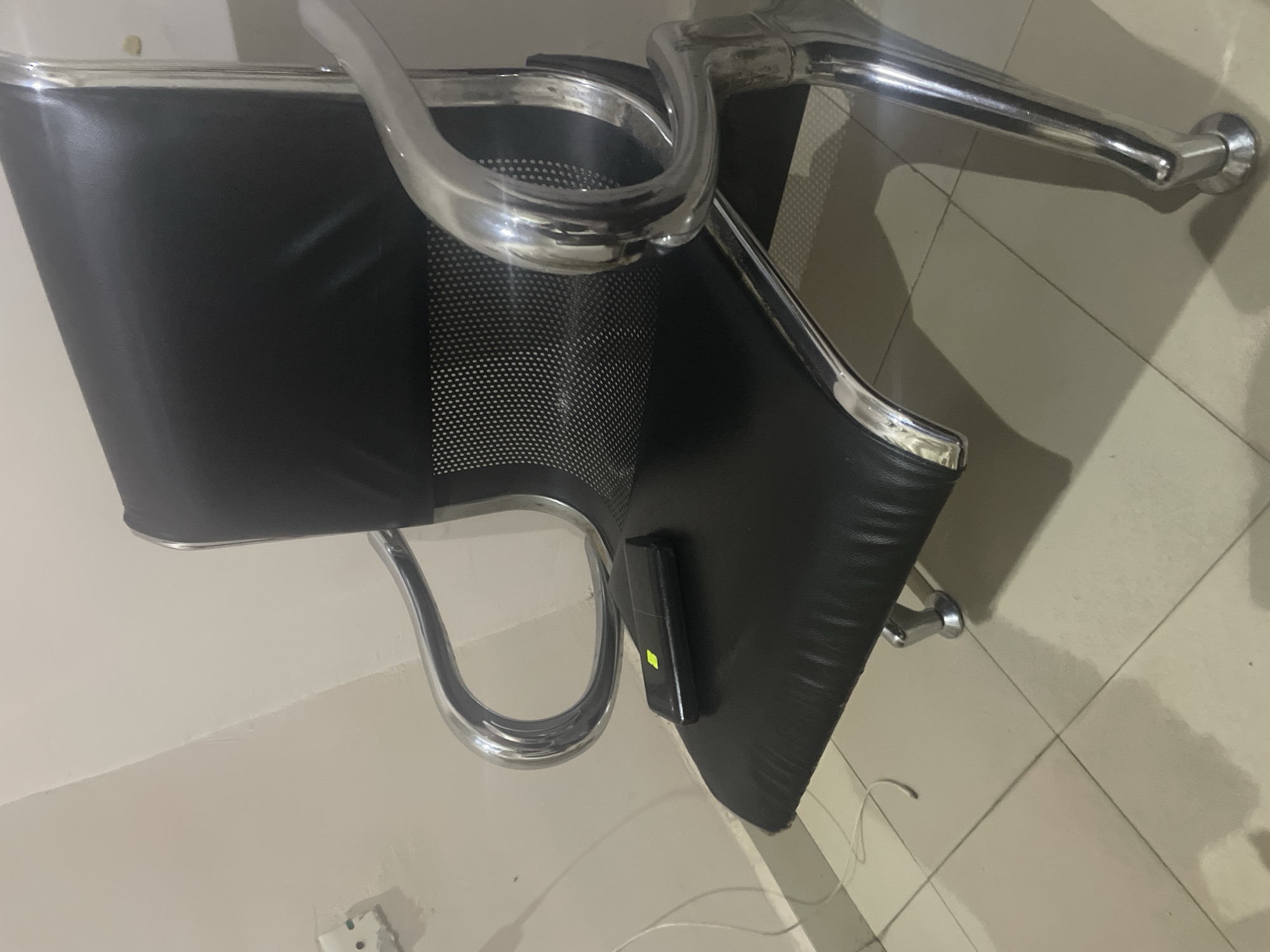 A black leather waiting chair with chrome legs sits on a tiled floor. A small black object with a green light is on the seat of the chair. The wall behind the chair is painted a light beige color. A white electrical outlet is visible on the wall. The scene appears to be indoors, likely in a waiting area or office. The location is specified as the city of Jalingo, Nigeria.