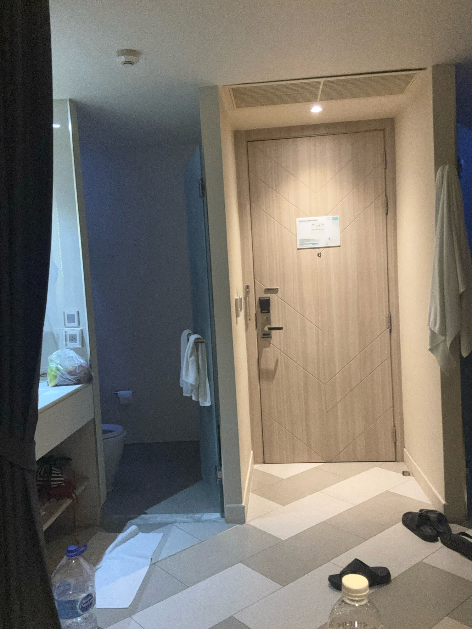 The image shows an interior view of a hotel room in Karon, Thailand. The room features a doorway with a light wood-grain finish and a digital lock. Above the door, a ventilation grate is visible on the ceiling. To the left of the doorway, an open bathroom area is partially visible, containing a toilet and a sink with a plastic bag of items on its surface. A towel hangs on a rack in the bathroom. The floor of the room is tiled with a pattern of light and dark grey squares. In the foreground, a large plastic bottle of water and a pair of black flip-flops are present on the floor. A dark curtain is visible on the far left side of the image. A smoke detector is mounted on the ceiling in the upper left corner.
