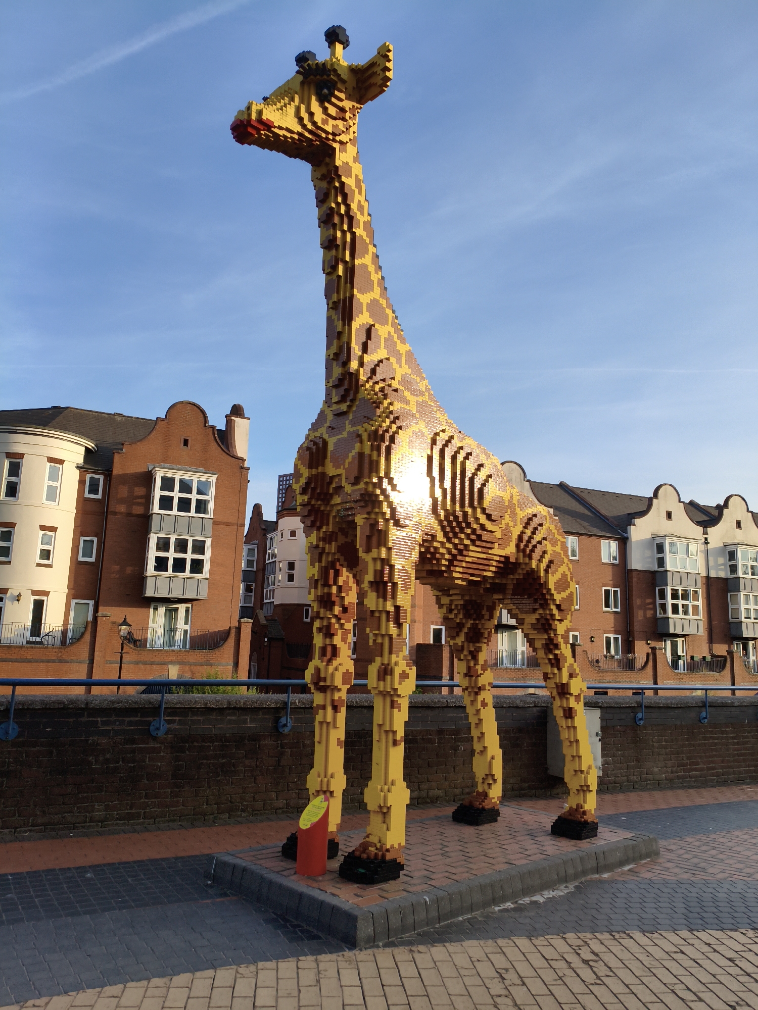 A tall, bright yellow and brown Lego giraffe sculpture stands on a paved walkway in the city of Birmingham, United Kingdom. The giraffe is constructed from numerous Lego bricks, with distinct brown patches on its yellow body. It has black Lego hooves and a red Lego muzzle. The sculpture is positioned in front of a row of red brick buildings with white-framed windows and a dark tiled roof. A blue railing runs along the edge of the walkway, separating it from a brick wall. The sky above is clear and blue, with faint white clouds. The sun is shining brightly, reflecting off the giraffe's body. The walkway is paved with interlocking bricks in shades of grey and orange.